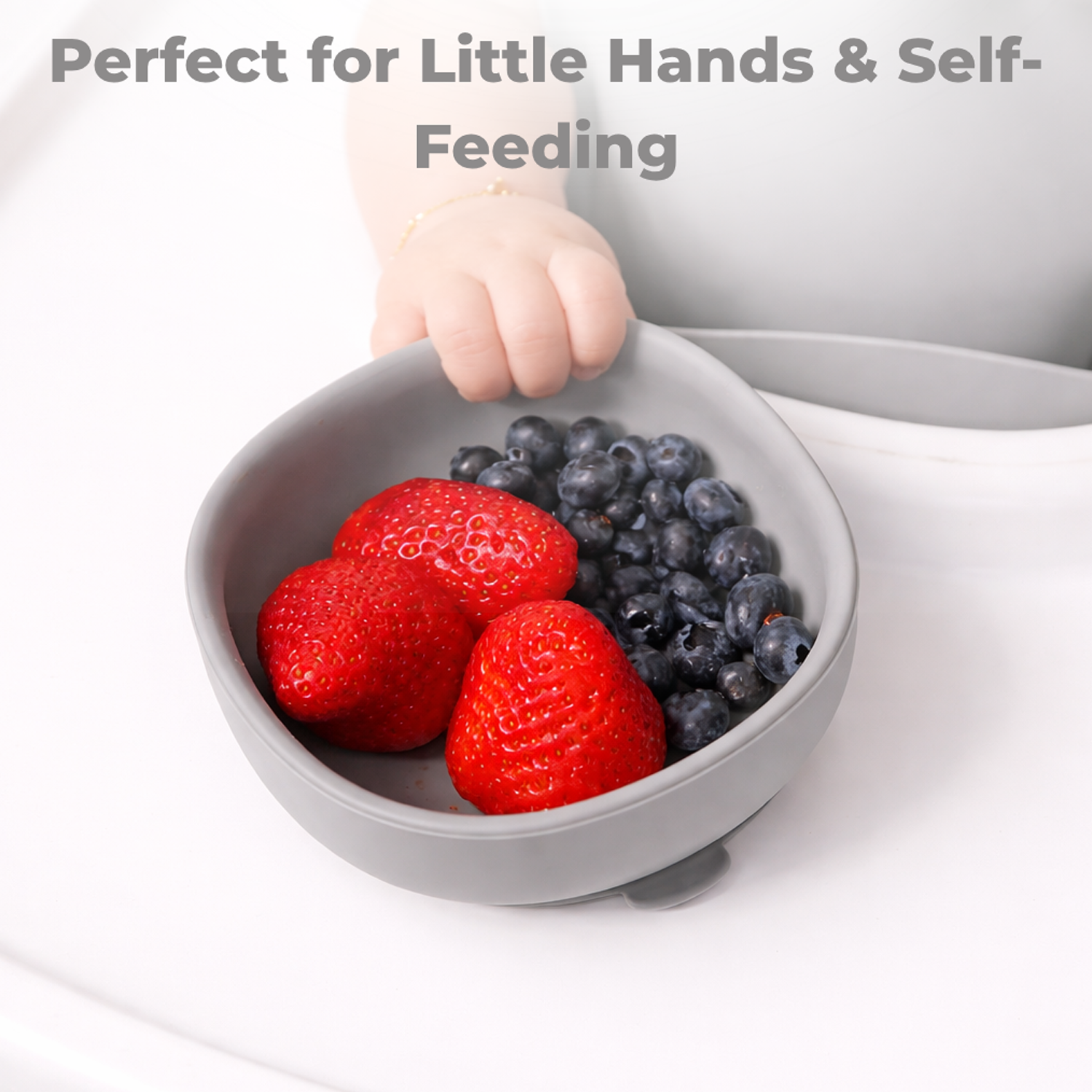 Gray silicone suction bowl with strawberries and blueberries held by a child's hand, text 'Perfect for Little Hands & Self-Feeding' above.