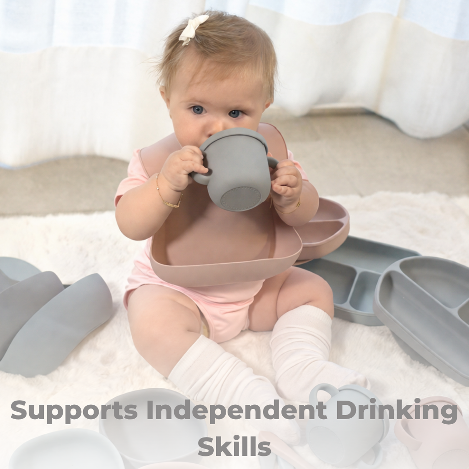 Baby sitting on a blanket holding a grey silicone training cup with a straw, surrounded by additional silicone products