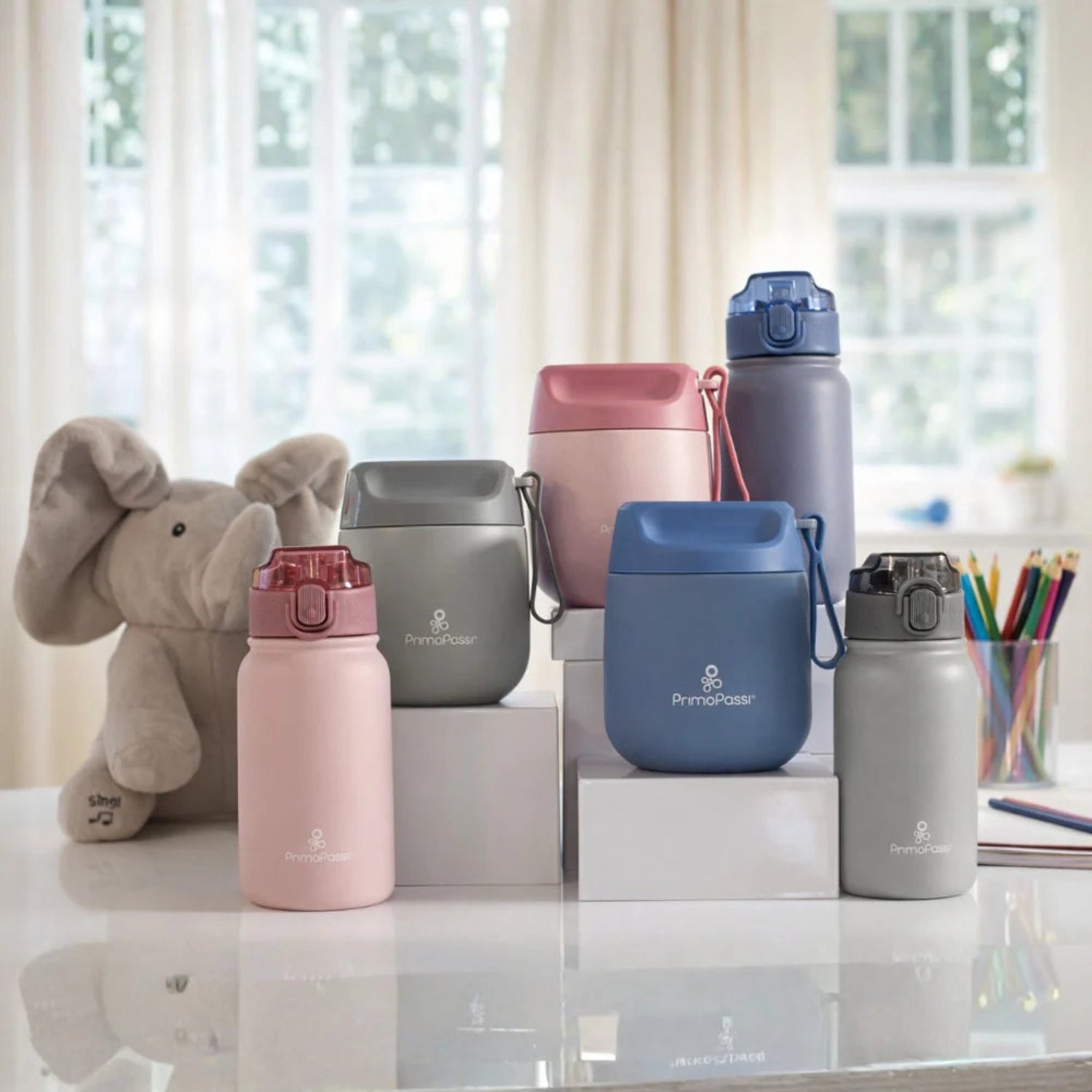 Collection of pink, grey and blue insulated food jars and water bottles on a reflective surface with a blurred window background.