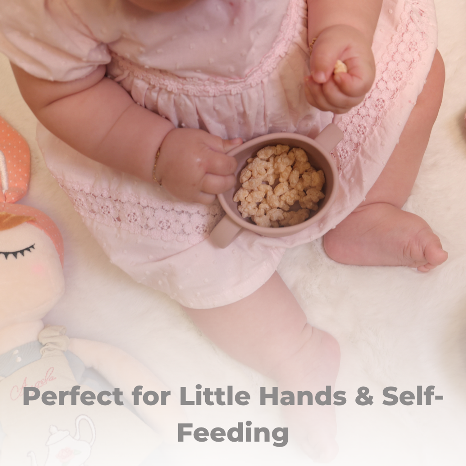 Baby in pink outfit holding a small cup with cereal, surrounded by soft toys on a light background