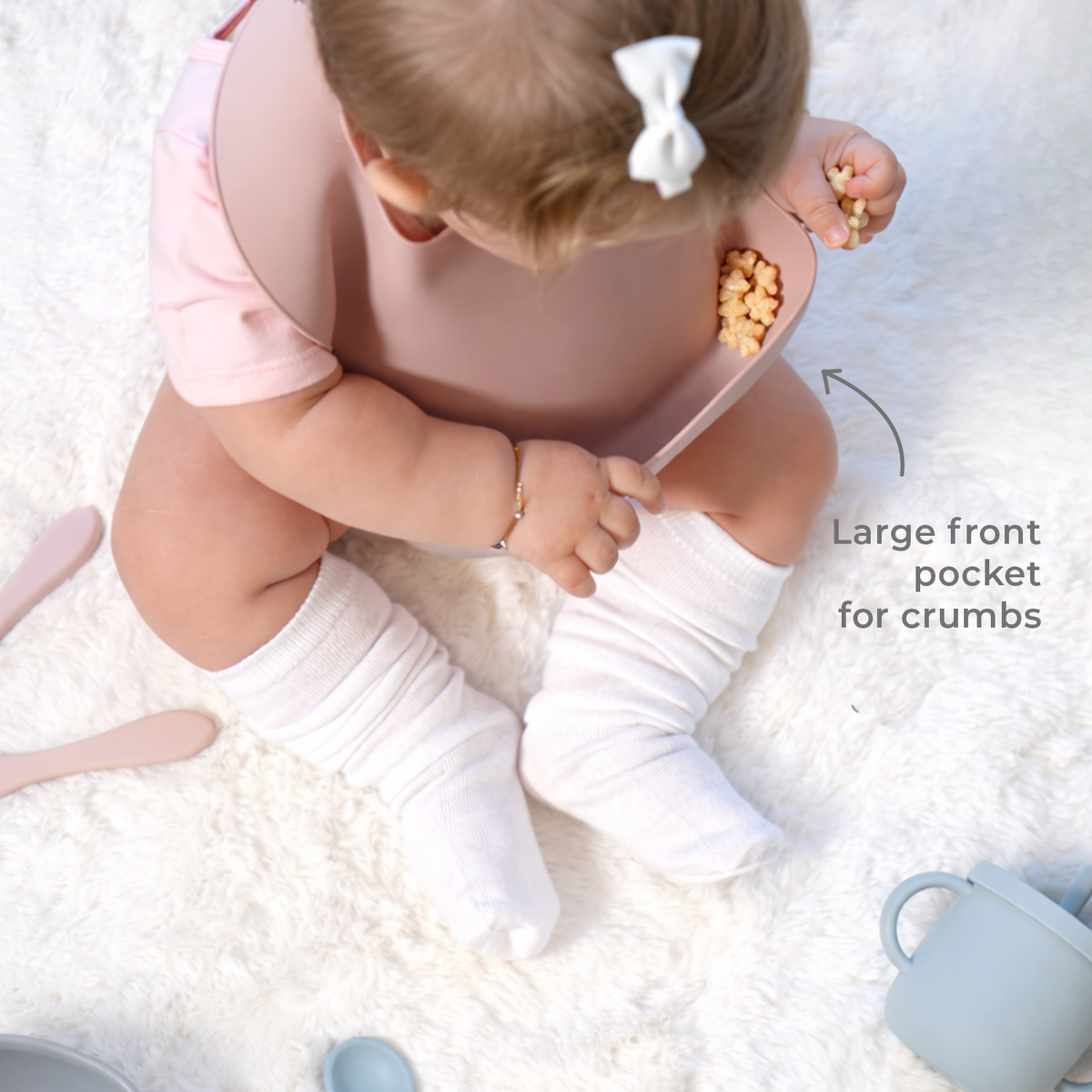 Baby sitting wearing a pink bib with a large front pocket for crumbs, surrounded by other silicone products on a white surface.
