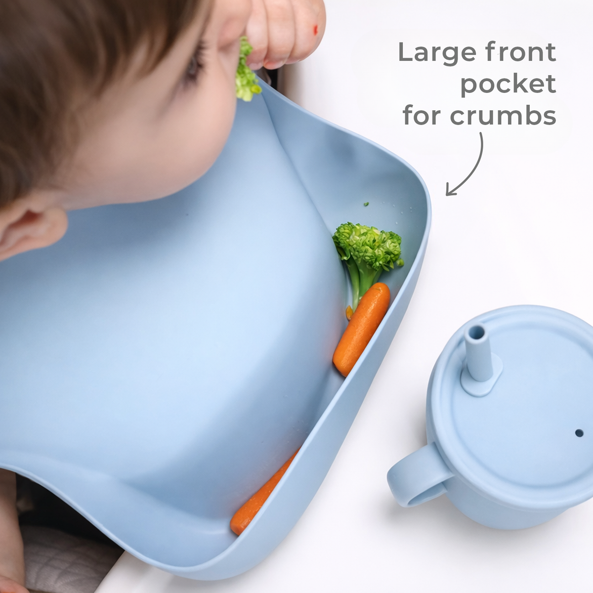 Child using a blue bib with a large front pocket for crumbs, containing vegetables and a cup.
