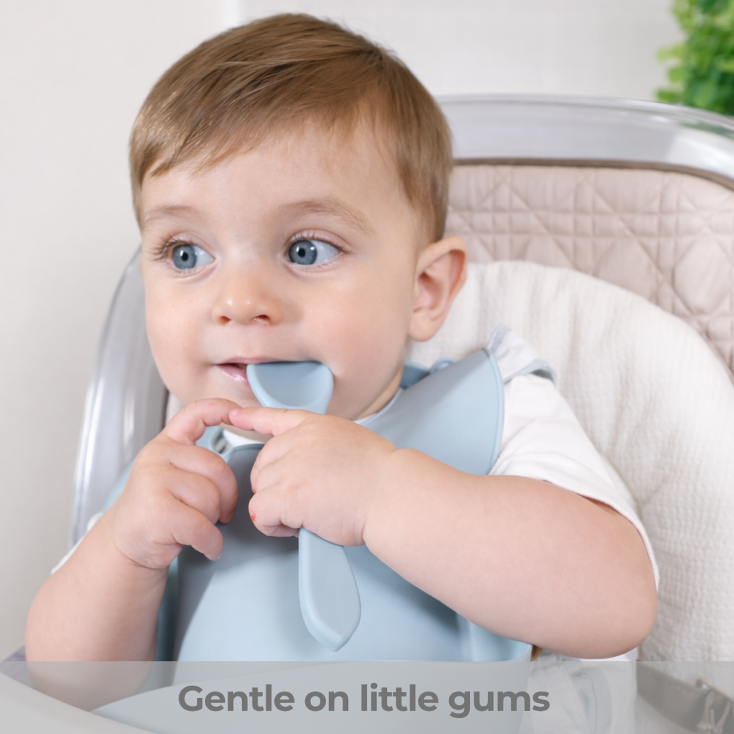 Baby sitting in a high chair with a blue silicone spoon in his mouth, wearing a bib. text: Gentle on little gums"