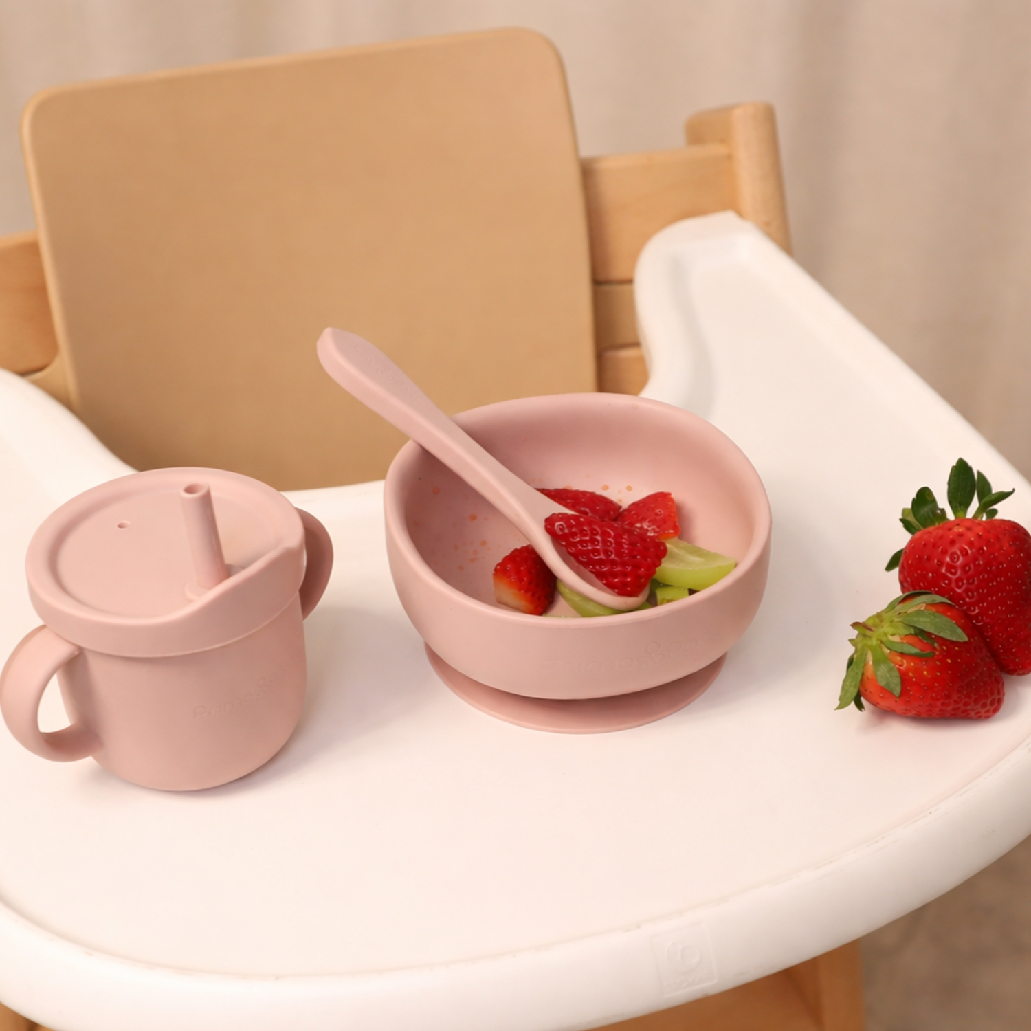 Pink suctiob bowl with a spoon and strawberries on a high chair, next to a pink sippy cup.