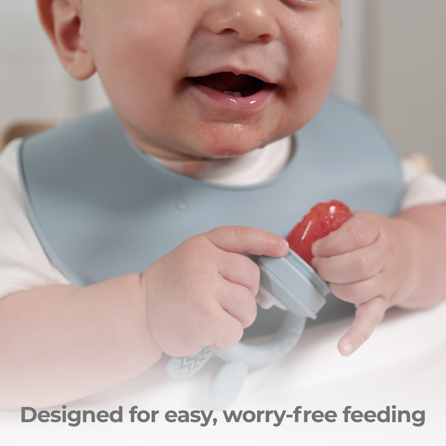 Baby wearing a bib holding a food feeder with strawberries on it with text 'Designed for easy, worry-free feeding'.