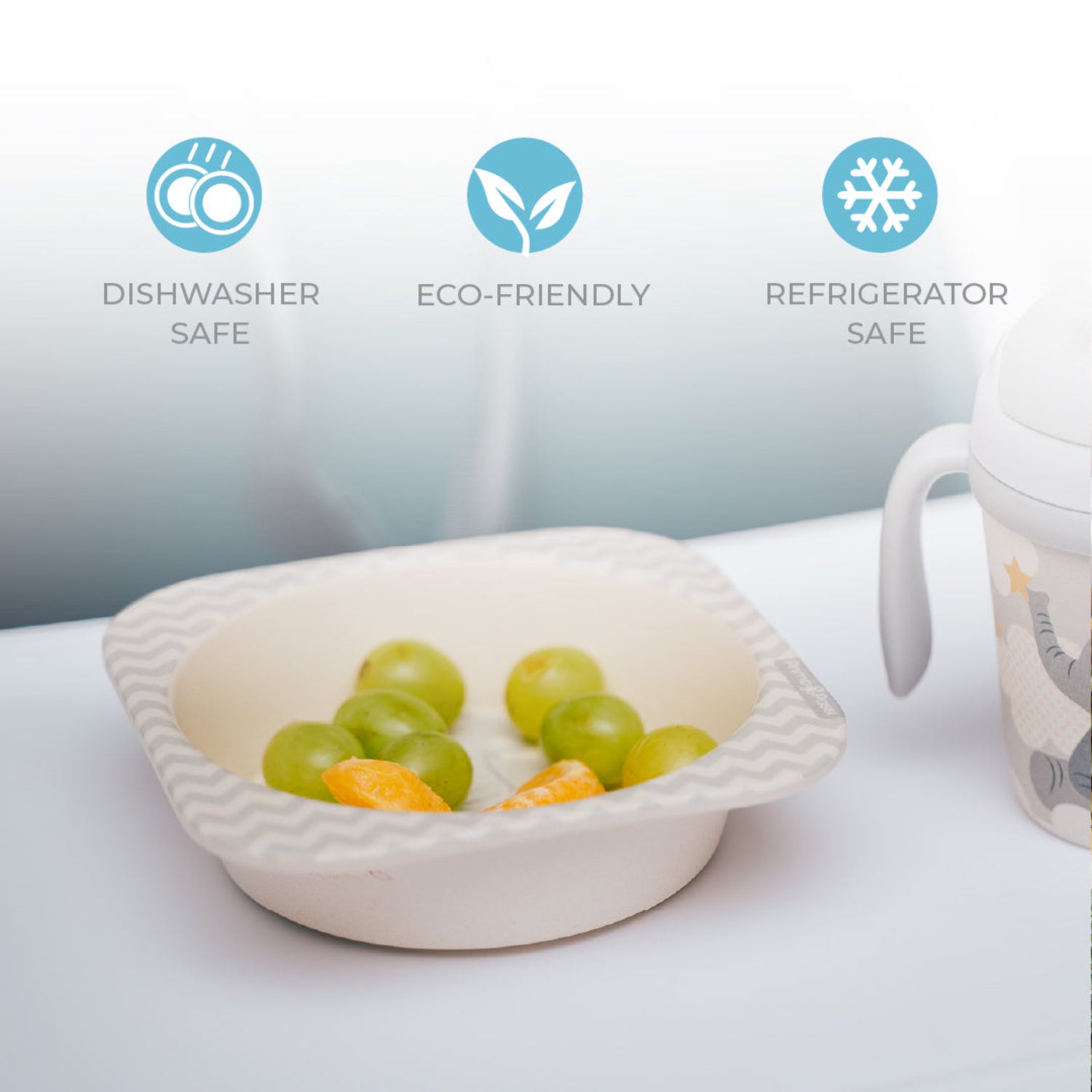 A bamboo fiber children's bowl filled with grapes and orange slices sits beside a matching elephant-themed toddler training cup. The image highlights that the bowl is dishwasher safe, eco-friendly, and refrigerator safe, making it ideal for everyday snacks and meals.