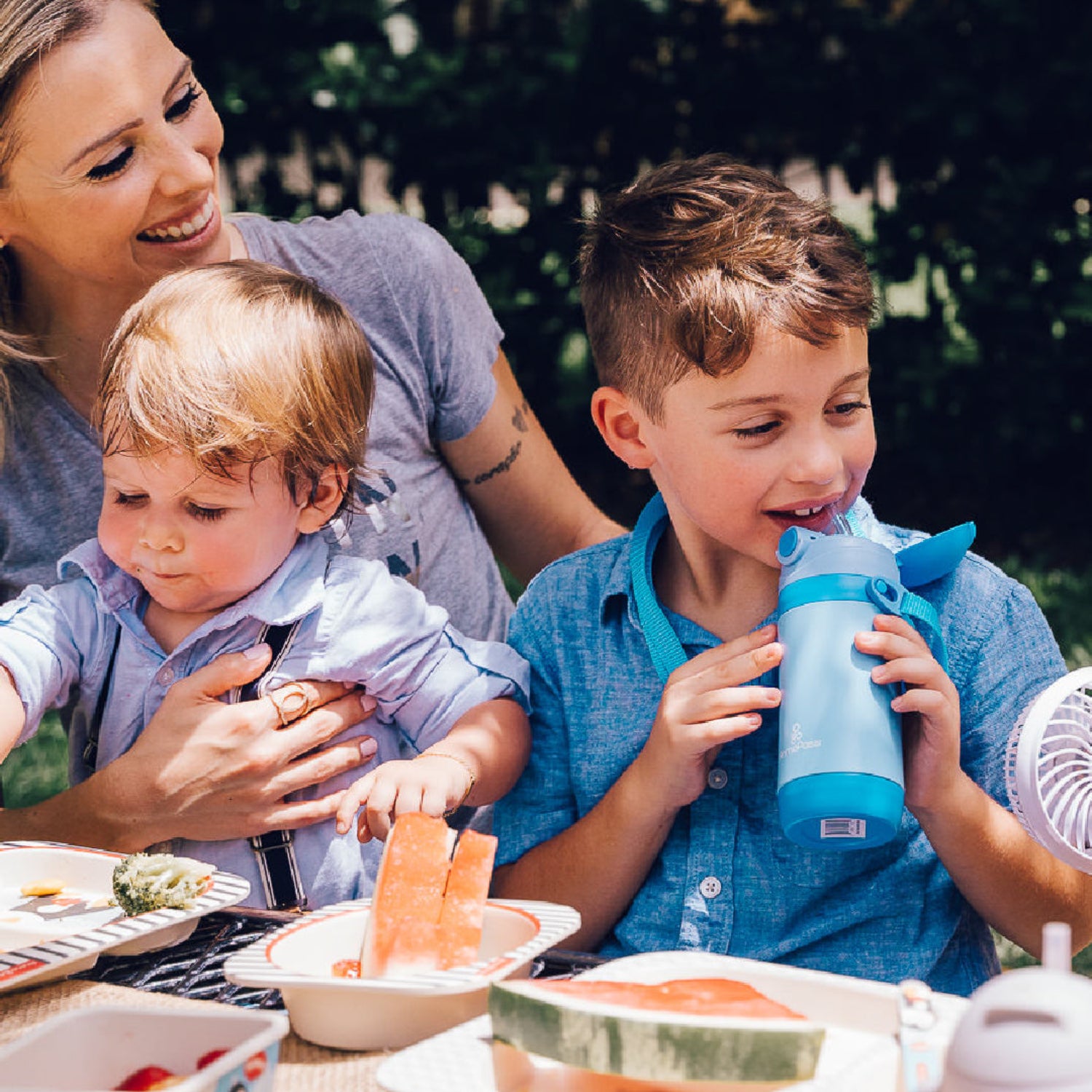 A mother enjoys an outdoor meal with two young children as one child drinks from a blue insulated straw bottle and the other eats from a bamboo fiber bowl. The lightweight, eco-friendly tableware makes family meals and picnics easy, safe, and fun.