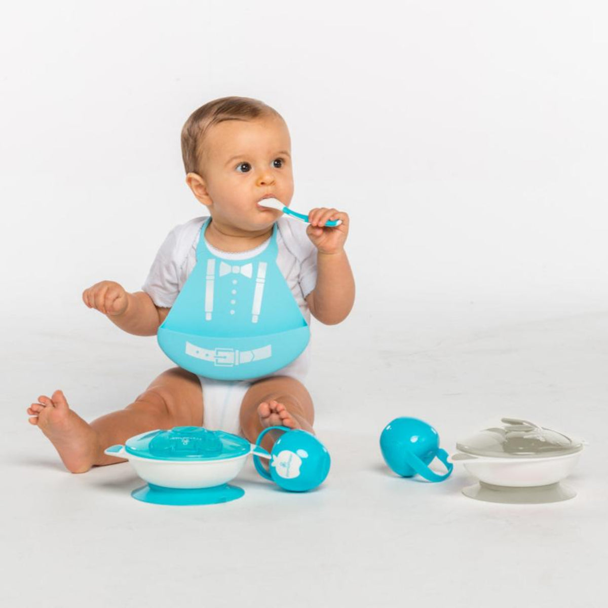 Baby wearing a turquoise Primo Passi silicone bib sits on the floor while holding a baby spoon. Surrounding the child are matching Primo Passi feeding essentials, including suction bowls with lids and two pacifier cases in blue and gray. The coordinated feeding collection is designed to support clean, convenient mealtimes at home or on the go.