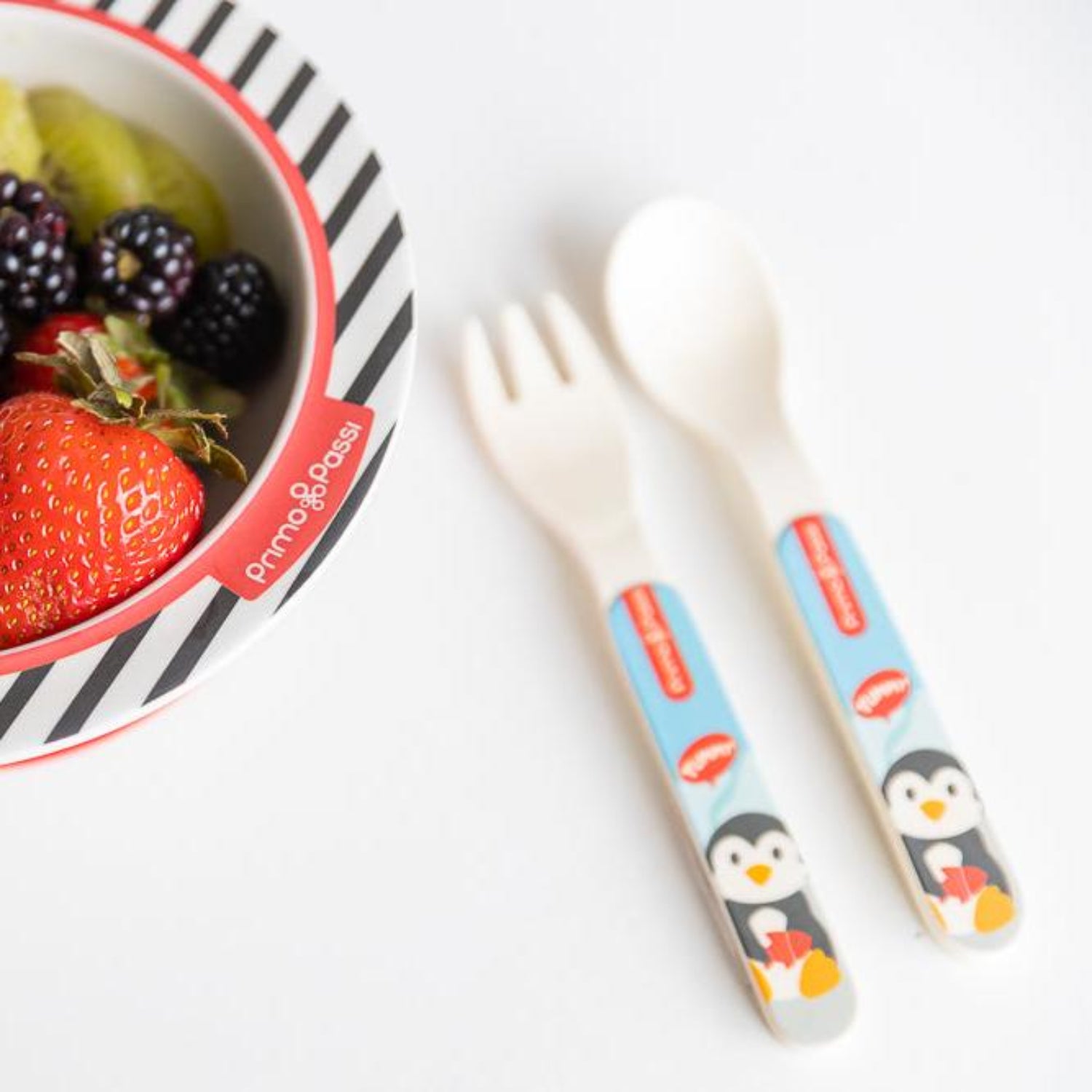 Toddler spoon and fork with colorful arctic-themed penguin handles are displayed beside a matching bamboo fiber bowl filled with fresh fruit, including strawberries, blackberries, and kiwi. The coordinated feeding set is designed for little hands learning to self-feed.