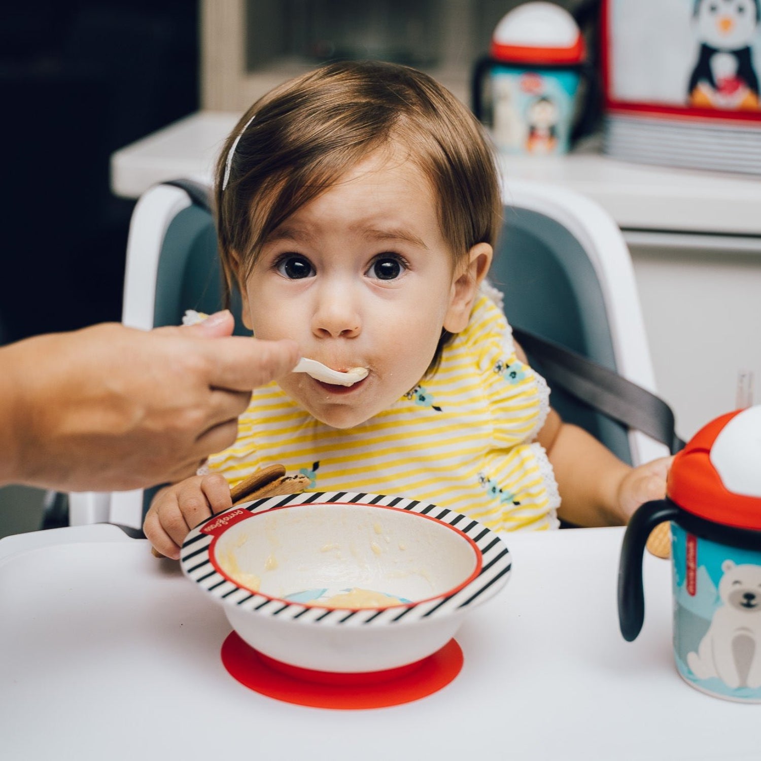 A baby in a yellow striped shirt sits in a high chair while an adult feeds them from a bowl. A colorful sippy cup is visible on the right side of the table.
