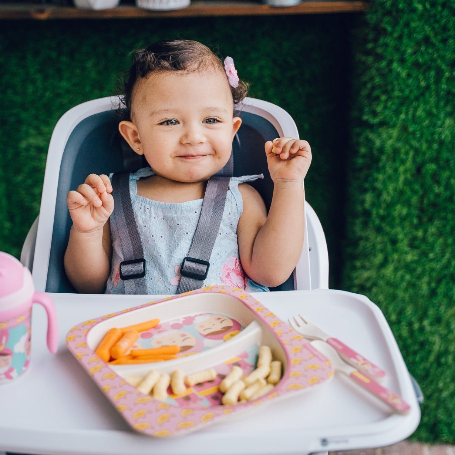 Toddler smiling in a high chair with a divided baby plate, training cup, fork, and spoon while enjoying a healthy self-feeding meal. The colorful feeding set encourages independent eating and makes mealtime fun.
