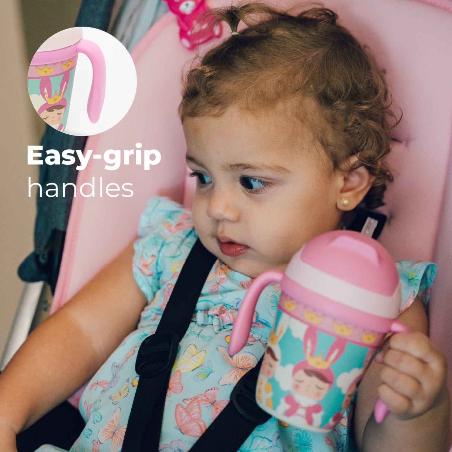 A little girl holding a Pink toddler training cup with two easy-grip handles, a soft straw, and a colorful princess-themed design featuring crowned characters, clouds, and crowns. With a text: easy-grip handles
