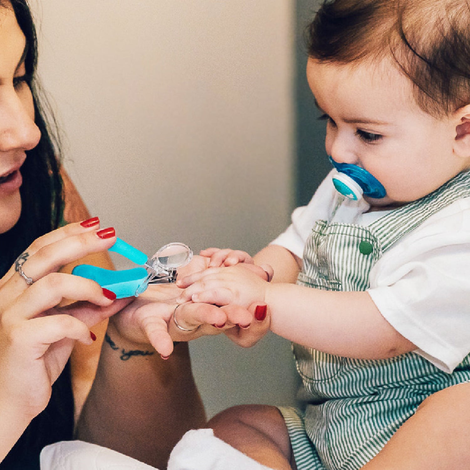 A baby wearing a green and white striped clothes and white t-shirt has their nails clipped by an adult while wearing a bright light blue nail clipper. The baby has a blue pacifier in their mouth.