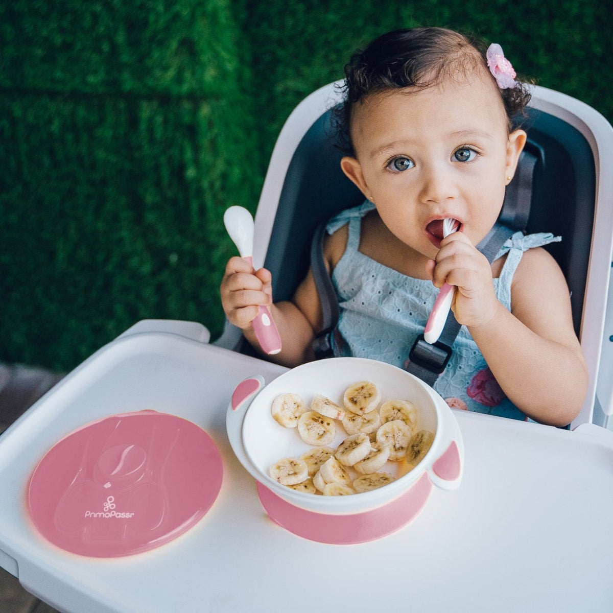 Toddler seated in a high chair using the Primo Passi suction bowl feeding set with sliced bananas. The child holds the matching spoon and fork while the white bowl with a pink silicone suction base stays securely in place. The pink lid with built-in utensil storage rests beside the bowl, showcasing a practical mealtime solution for babies learning to self-feed.