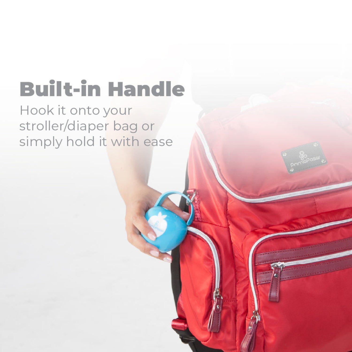 Person holding the blue Primo Passi pacifier case attached to a red diaper backpack using its built-in flexible handle. The compact case securely stores pacifiers, keeping them clean and easily accessible while attached to a stroller, diaper bag, or carried by hand for everyday outings.