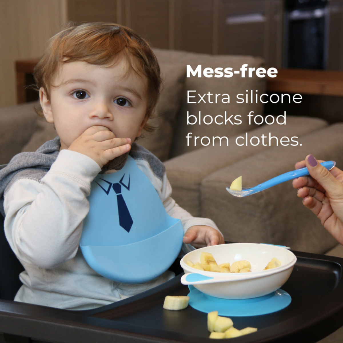 Toddler sitting in a high chair wearing a blue Primo Passi silicone bib with a tuxedo design while enjoying a snack. A white suction bowl with sliced bananas is securely attached to the tray, and an adult offers food with a matching spoon. The image highlights the bib's deep catch pocket for mess-free mealtimes, helping keep food off clothing.