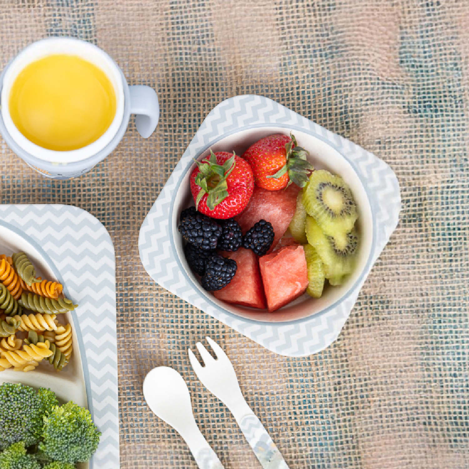 Top-down view of a bamboo fiber children's bowl filled with fresh fruit, including strawberries, blackberries, kiwi, and watermelon. A matching divided plate with pasta and broccoli, toddler spoon and fork, and a cup complete the coordinated elephant-themed mealtime set.