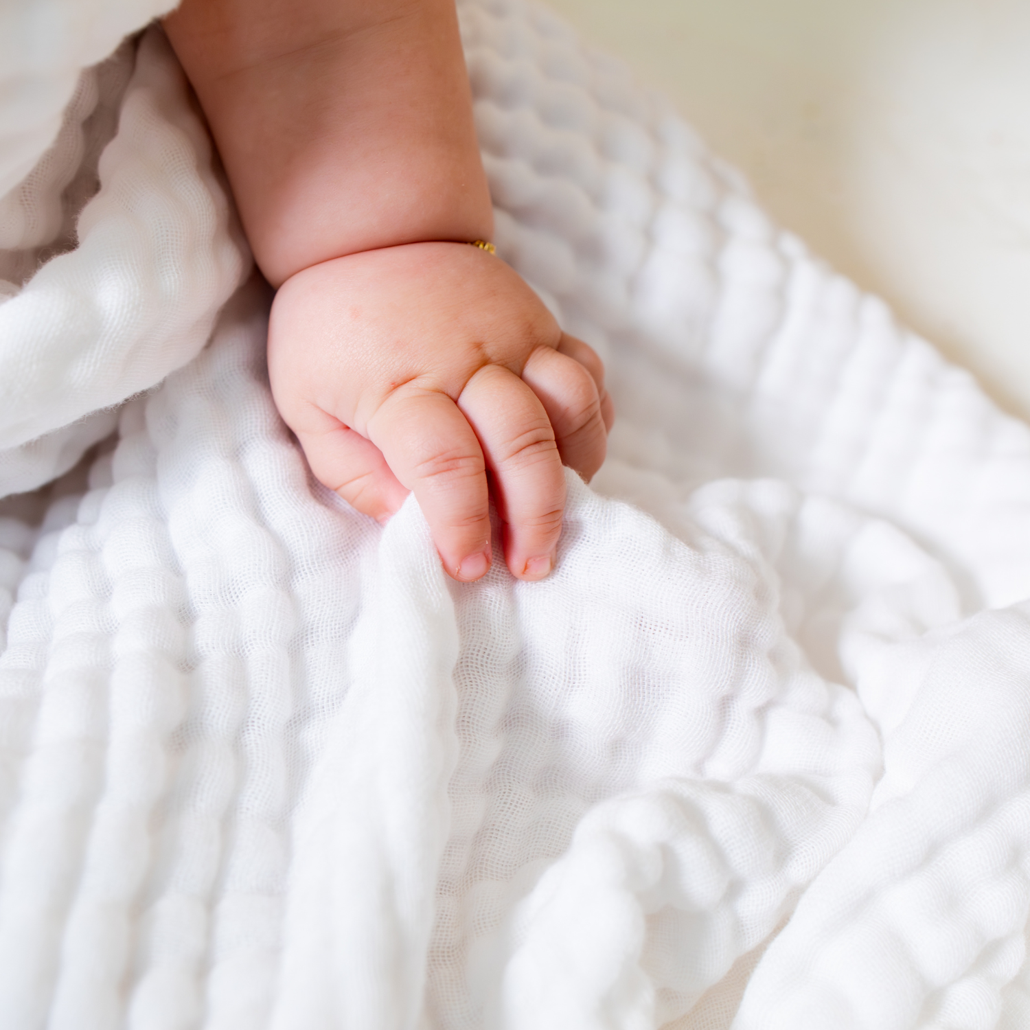 A close-up of a baby's hand resting on a soft, textured white muslin towel, with a tiny gold ring on the wrist.
