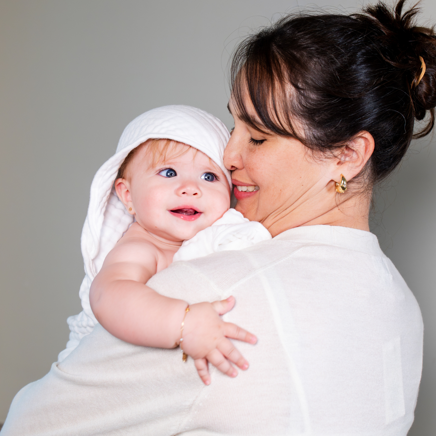 A smiling woman in a white top holds a baby wrapped in a white muslin towel. The background is a solid neutral gray studio backdrop.