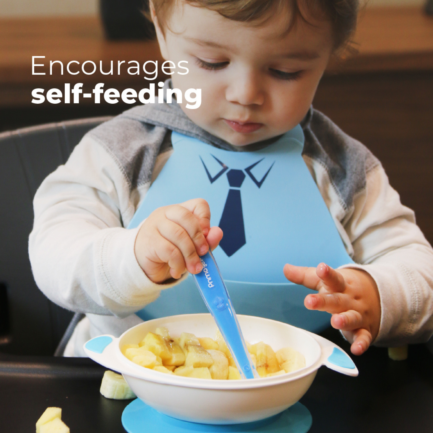 A toddler wearing a light blue bib with a black tie graphic sits in a high chair, using a blue-handled spoon to eat fruit from a white bowl. White text in the upper left reads 'Encourages self-feeding'.