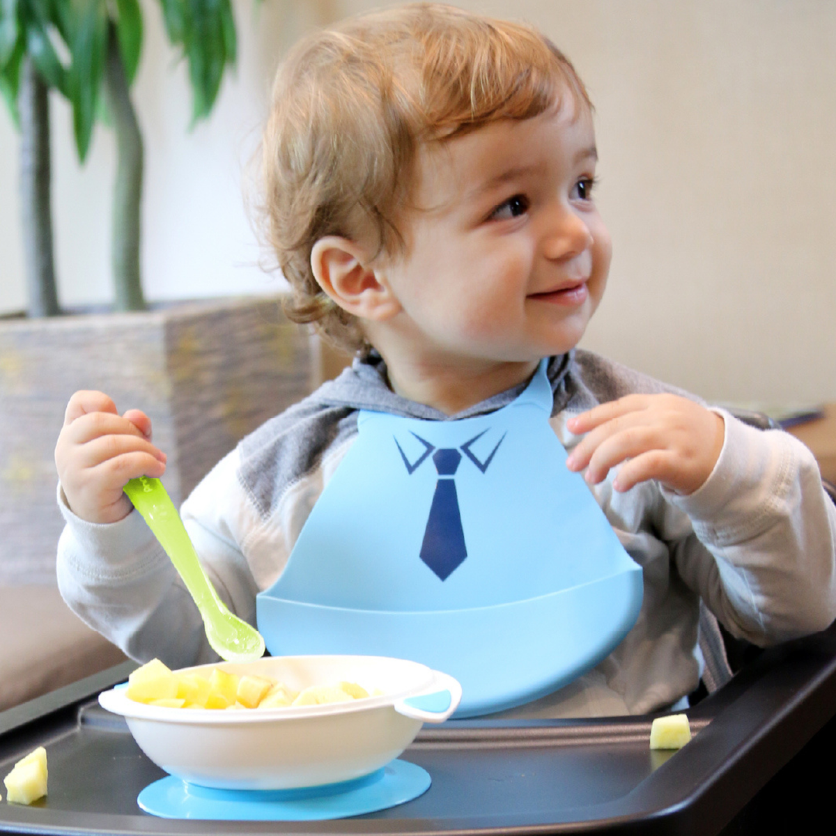 A baby wearing a light blue bib with a printed navy tie design sits in a high chair, holding a green spoon over a bowl of yellow fruit pieces.