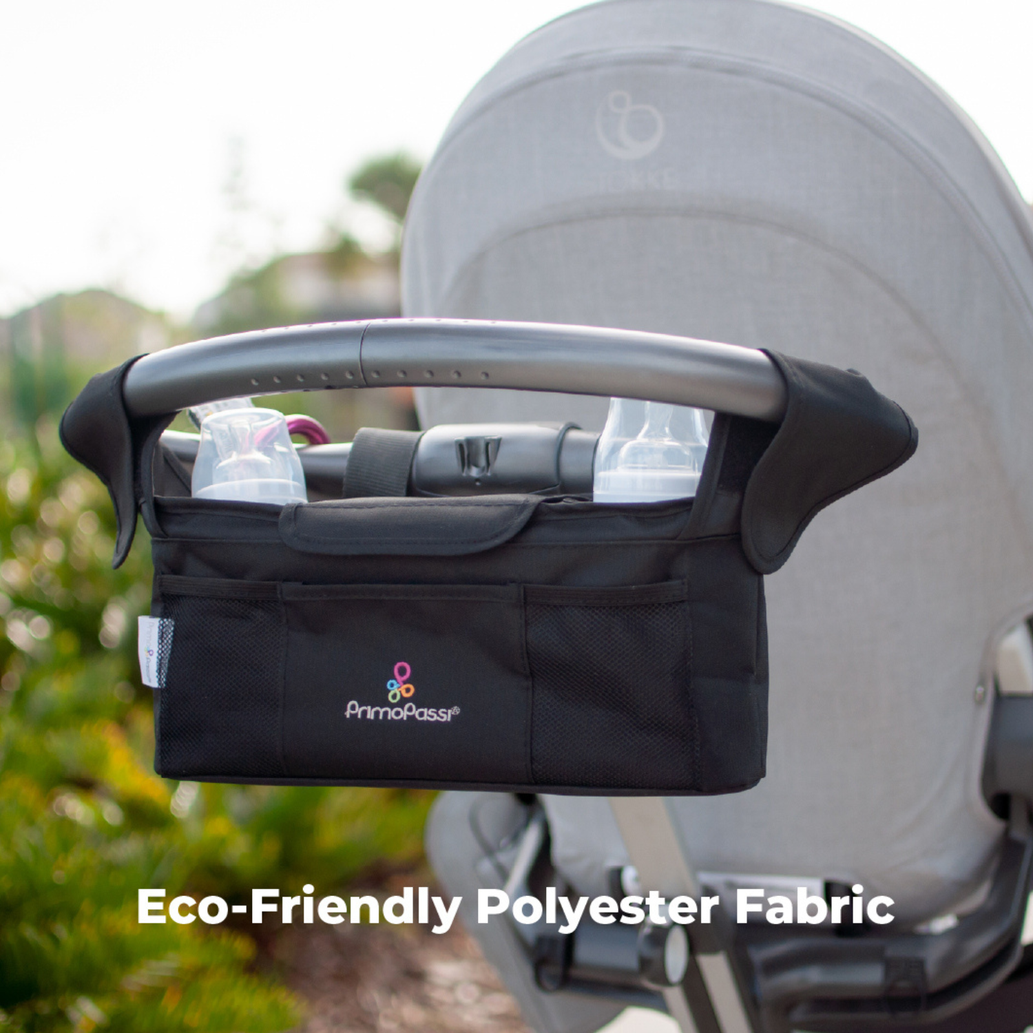 Primo Passi stroller handle organizer attached to a stroller outdoors, made from durable eco-friendly polyester fabric. The organizer features two insulated bottle holders, a spacious center compartment with magnetic closure, front and mesh storage pockets, and adjustable straps for secure attachment, keeping baby essentials organized and within easy reach during walks.