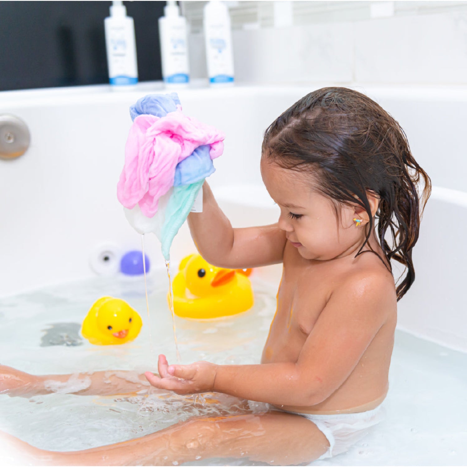 A young child plays in the bathtub while holding colorful muslin washcloths, squeezing out water during bath time. The soft, lightweight washcloths are gentle on delicate skin and become softer with every wash.