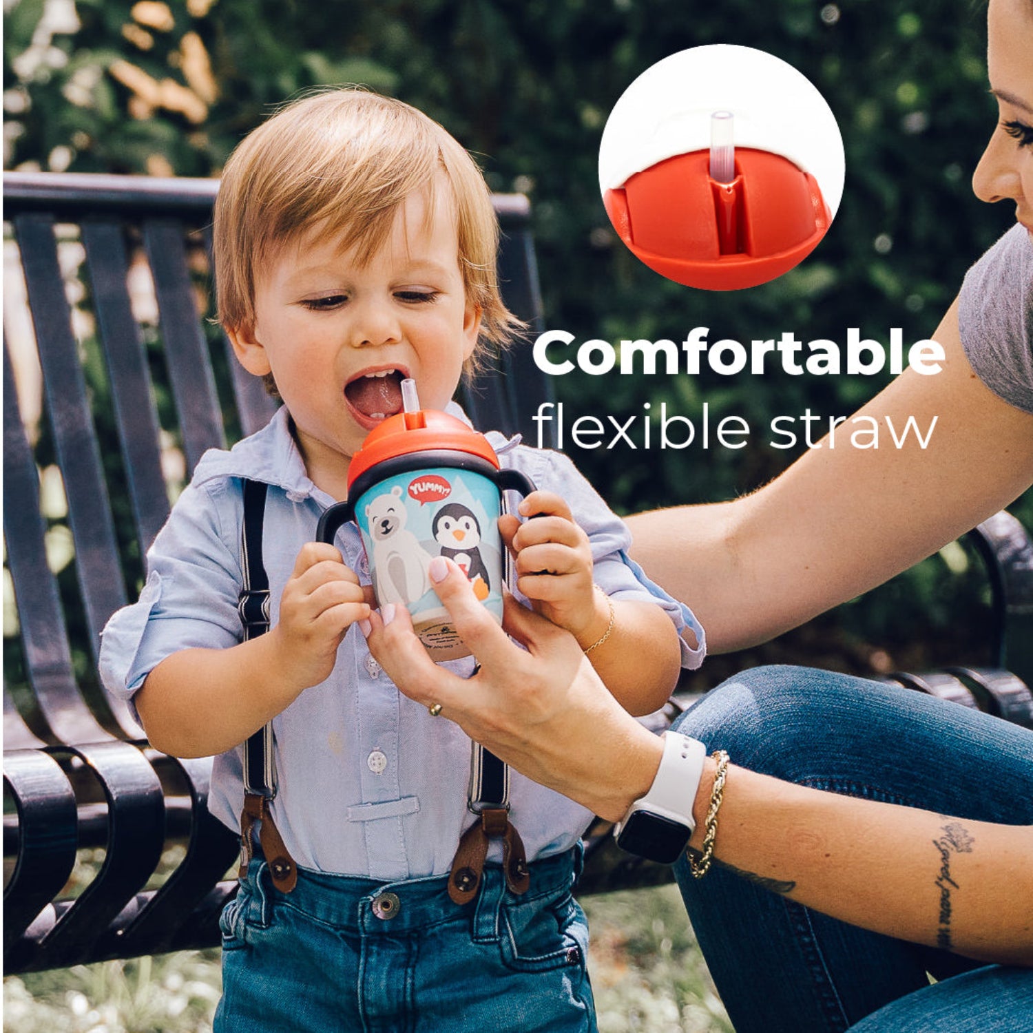 A little boy holding a Red toddler training cup with two easy-grip handles, a soft straw, and a playful arctic-themed design featuring a smiling polar bear and penguin against snowy mountains in a park background