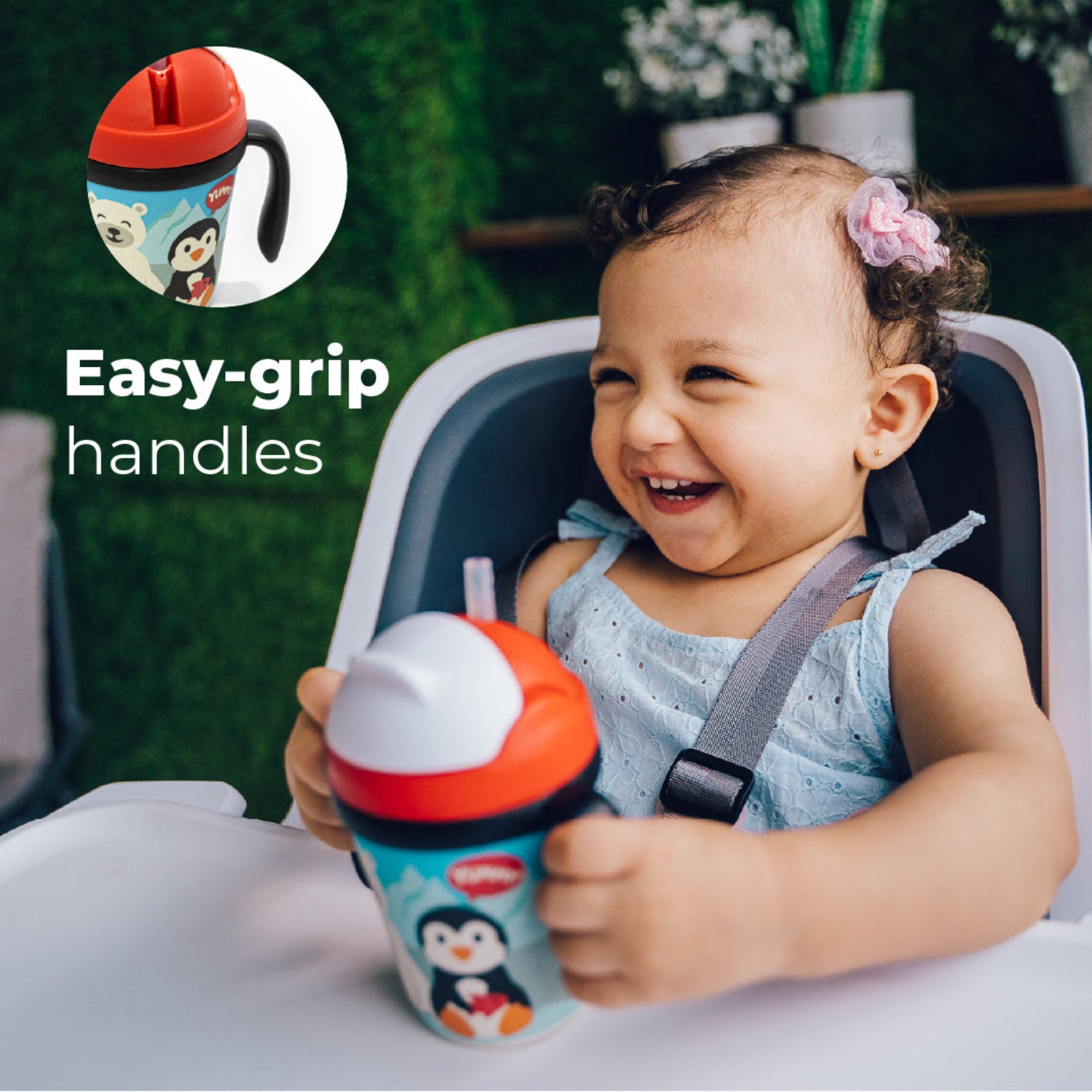 A little girl in a highchair holding a Red toddler training cup with two easy-grip handles, a soft straw, and a playful arctic-themed design featuring a smiling polar bear and penguin against snowy mountains. The spill-resistant cup is shown on a white background. with a text: easy-grip handles