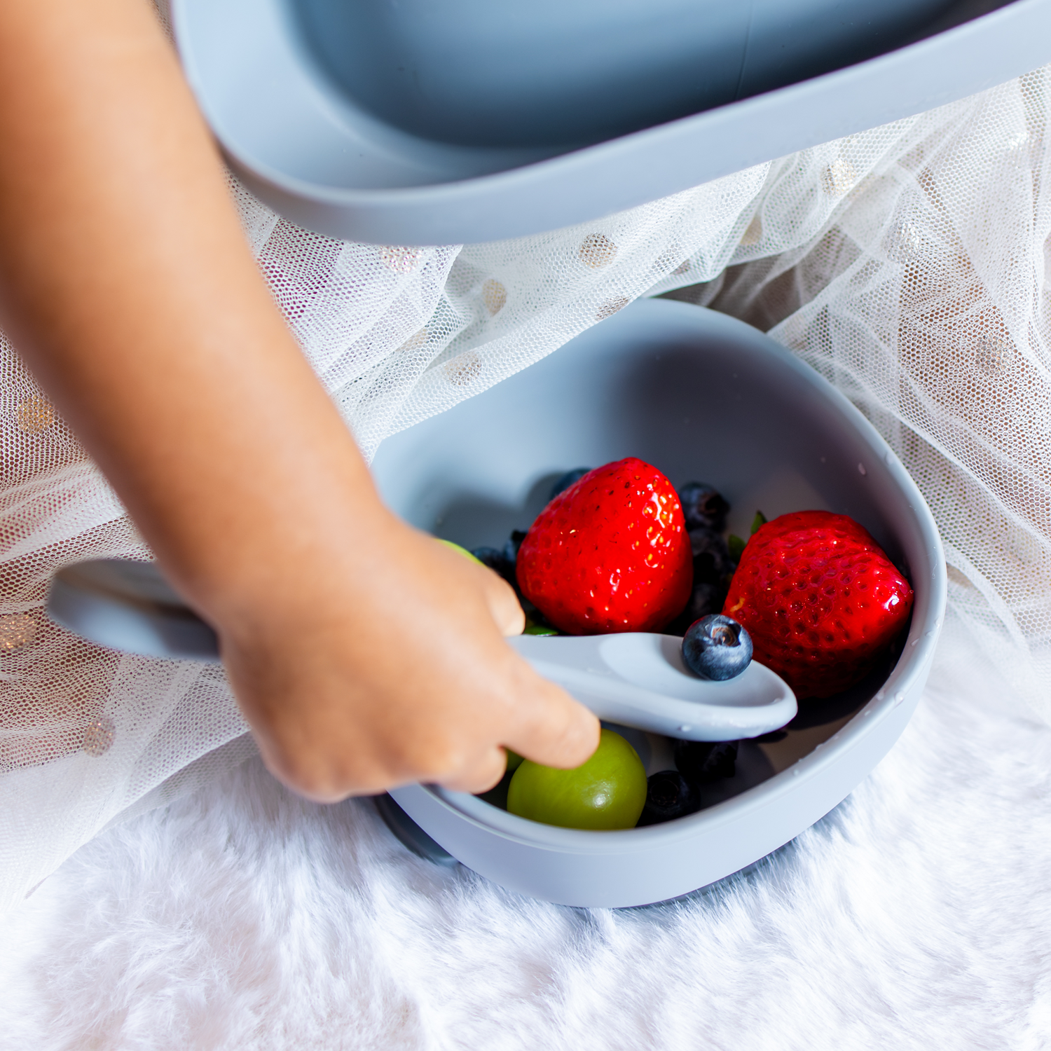 Child's hand reaching into a blue silicone bowl filled with fruits on a white surface.