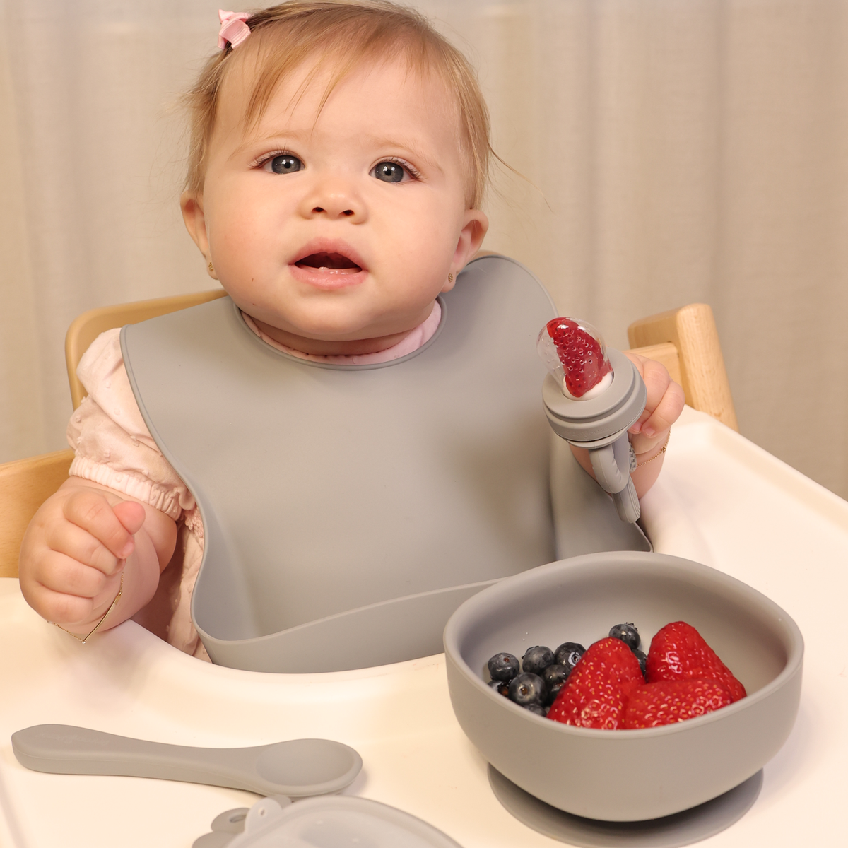 Baby sitting in a highchair with a bowl of fruits and wearing a grey bib, holding a food feeder with a strawberry on it.