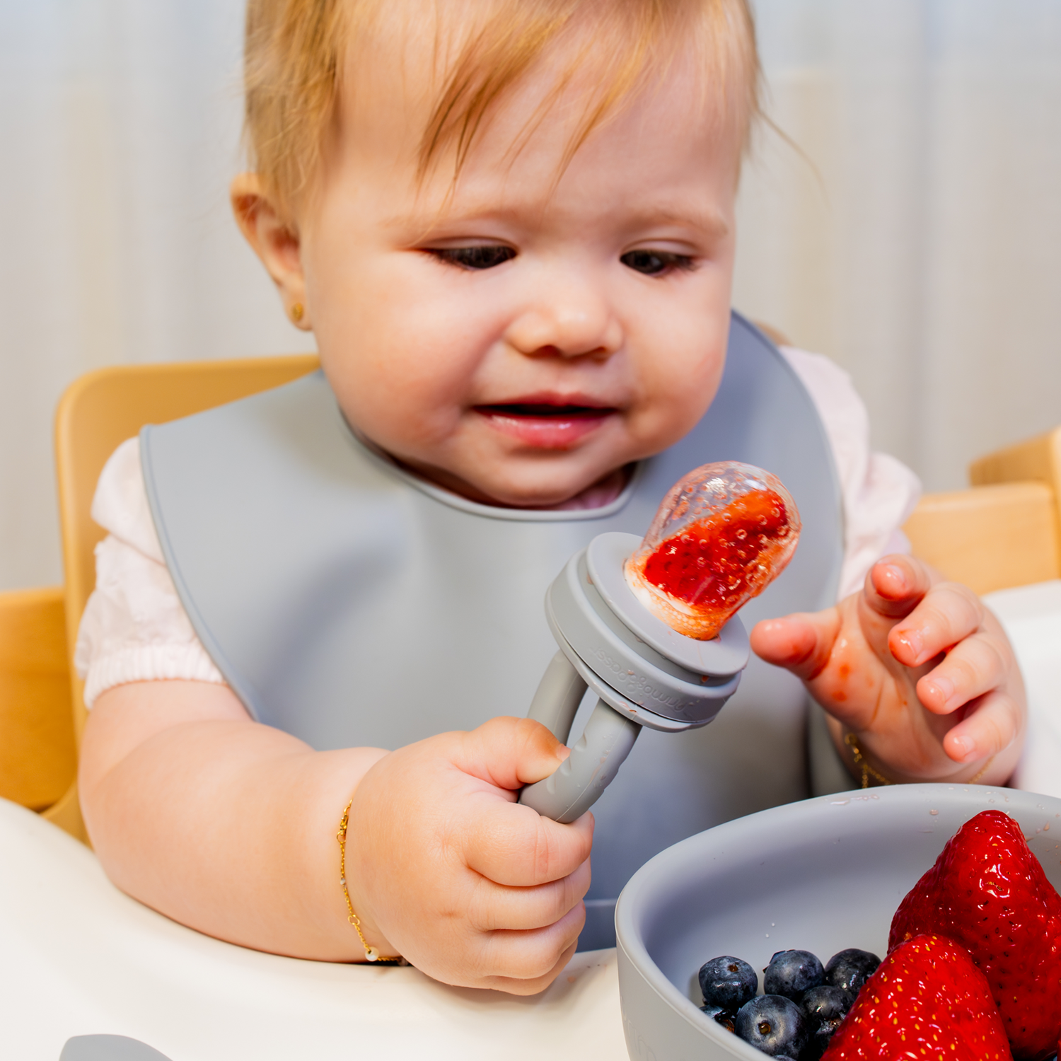 Baby in a high chair with a bib holding a food feeder with strawberry inside it, surrounded by fruits on a bowl.