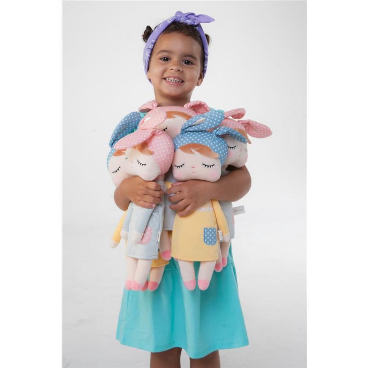 Smiling young girl wearing a turquoise dress and purple headband while hugging four soft plush dolls. The cuddly dolls feature embroidered closed eyes, colorful bow headbands, and pastel outfits, making them perfect companions for imaginative play, comfort, and snuggles.