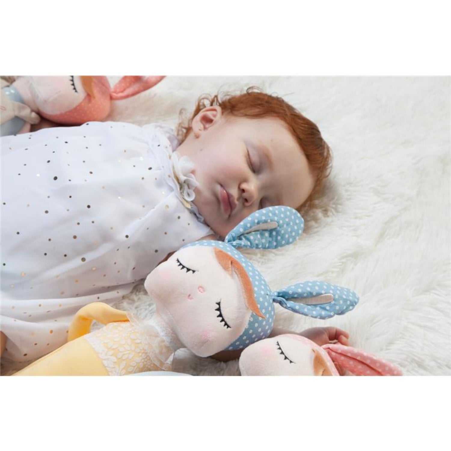 Sleeping toddler wrapped in a white swaddle blanket on a soft white rug, cuddling plush bunny dolls with embroidered closed eyes and pastel bunny ear headbands. The cozy scene highlights the dolls as comforting companions for naps, bedtime, and gentle snuggles.
