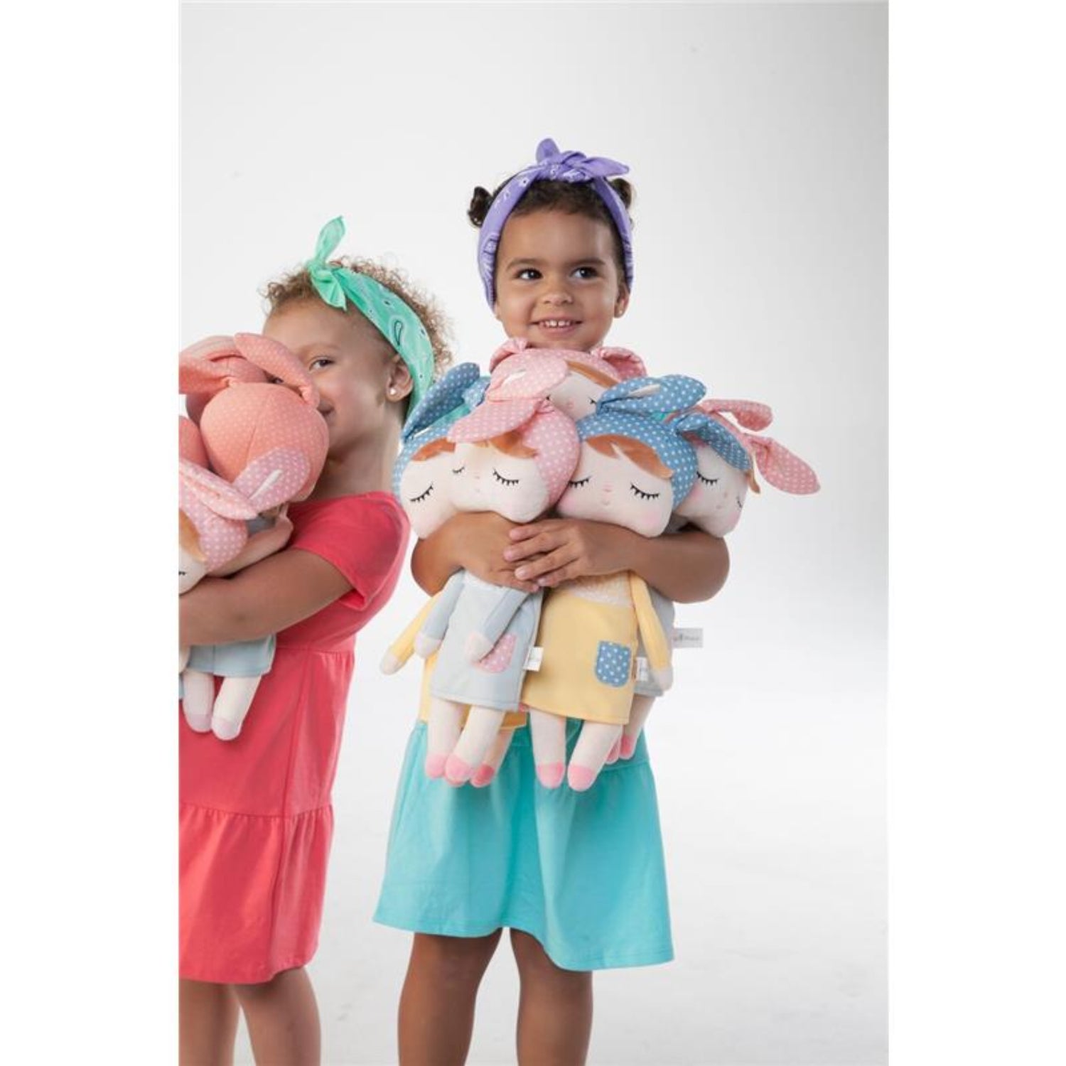 Two young girls wearing colorful dresses and headbands smile while hugging several soft plush bunny dolls. The pastel dolls feature embroidered closed eyes, floppy bunny ears, and cozy outfits, making them perfect companions for cuddles, imaginative play, and comforting moments.