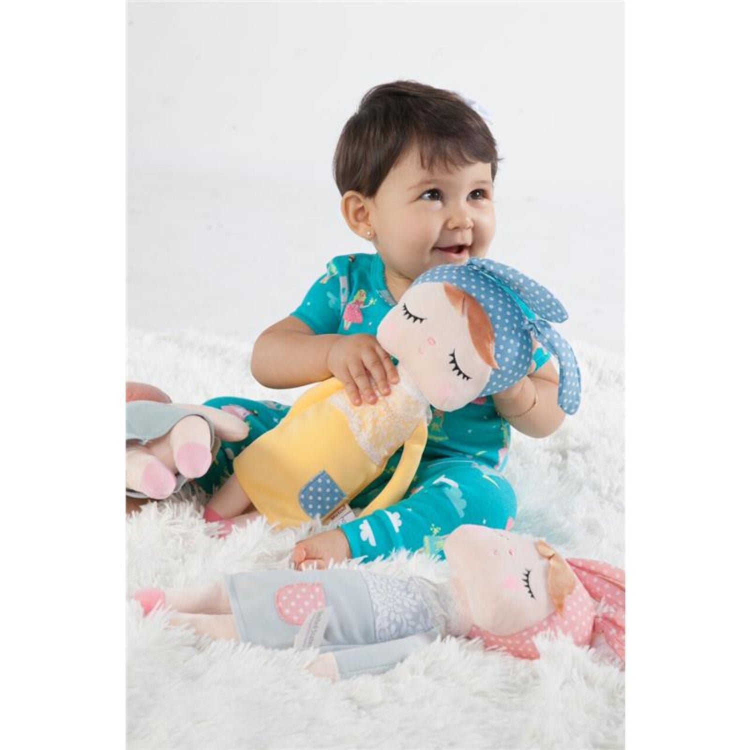 Toddler sitting on a soft white rug while cuddling a plush bunny doll with embroidered closed eyes, a blue polka-dot bunny ear headband, and a yellow dress. Additional pastel plush dolls surround the child, creating a warm, cozy setting for imaginative play, comfort, and snuggles.