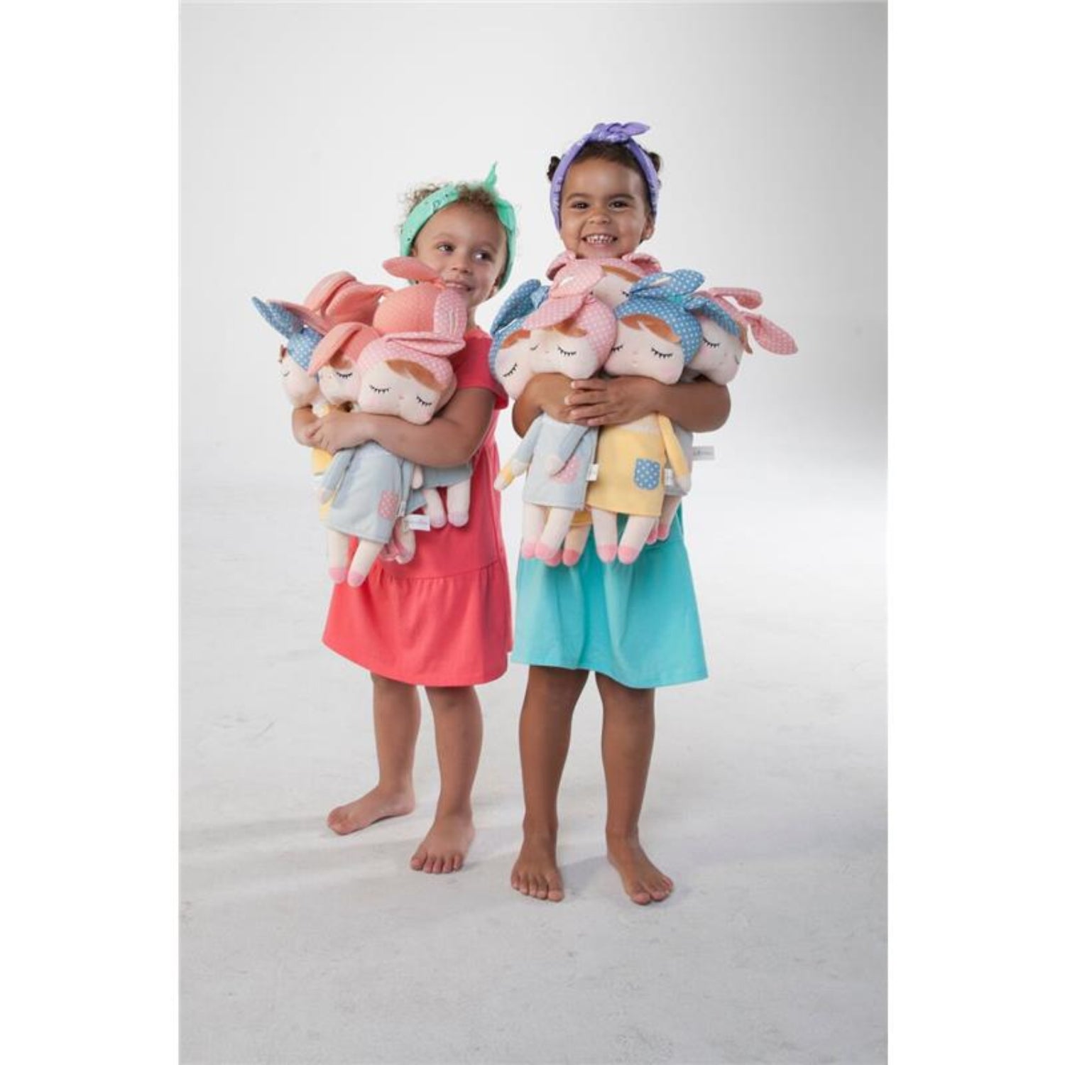 Two smiling young girls wearing colorful dresses and headbands while each hugs several soft plush dolls. The pastel dolls feature embroidered closed eyes, bow headbands, and cozy outfits, making them perfect for comforting hugs, imaginative play, and bedtime companionship.