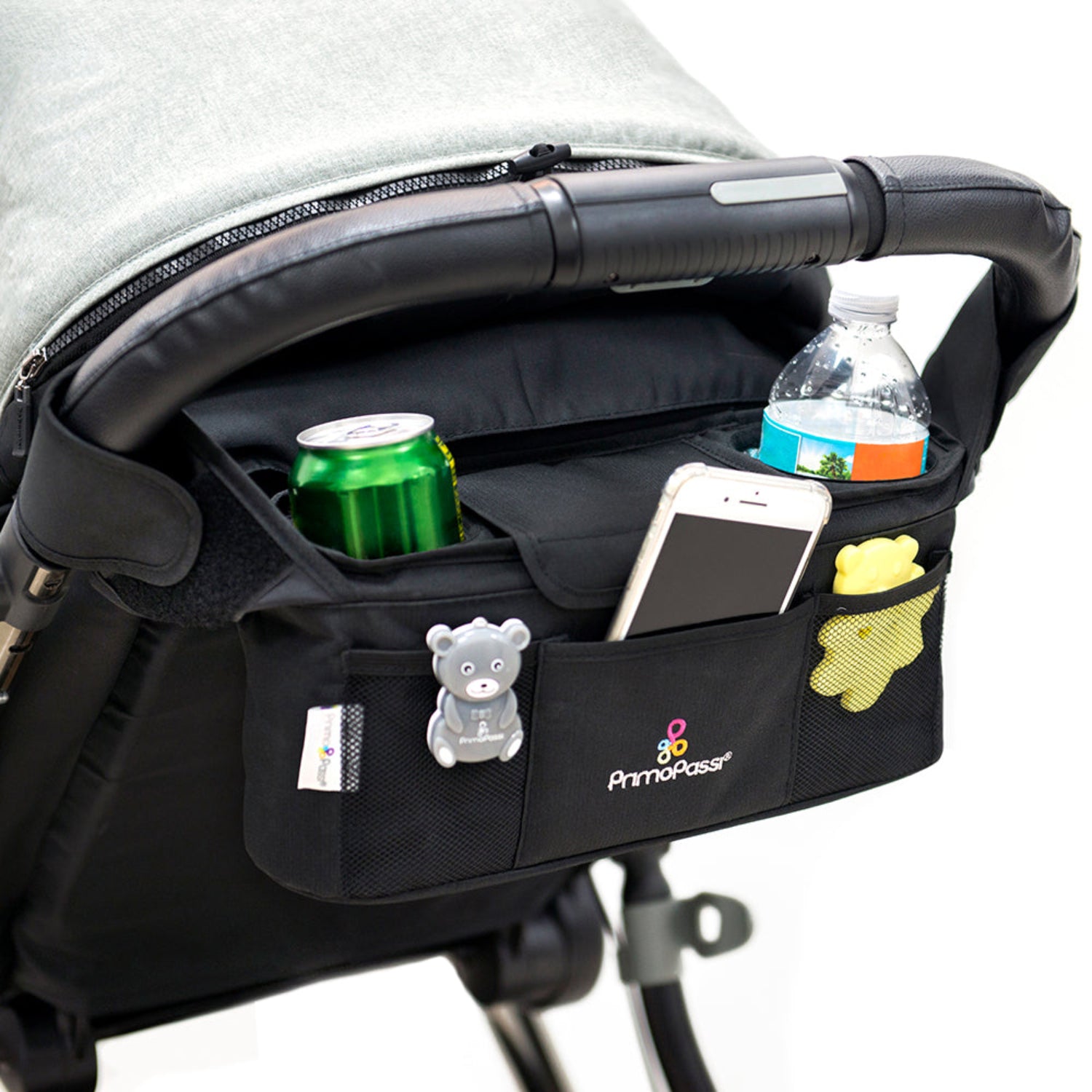 Primo Passi black stroller organizer attached to a stroller handle on a white background. The organizer features two insulated cup holders holding a water bottle and a soda can, a center compartment with a smartphone, front and side mesh pockets containing small baby essentials, and a bear-shaped stroller toy clipped to the front pocket, showcasing its spacious and organized storage design.