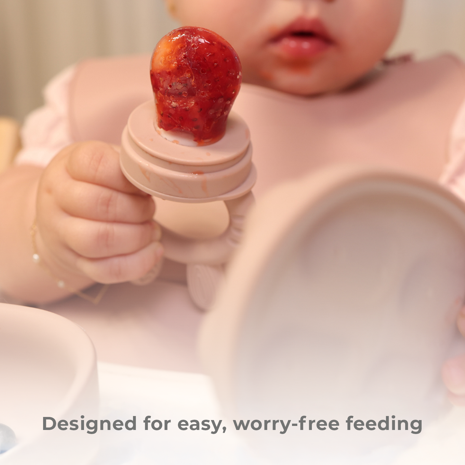 Baby holding a pink food feeder with a strawberry on it, designed for easy feeding.