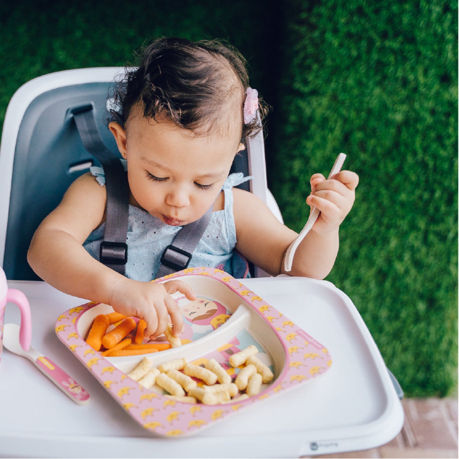 A young child with brown hair sitting in a high chair, eating carrots from a plate, holding a knife