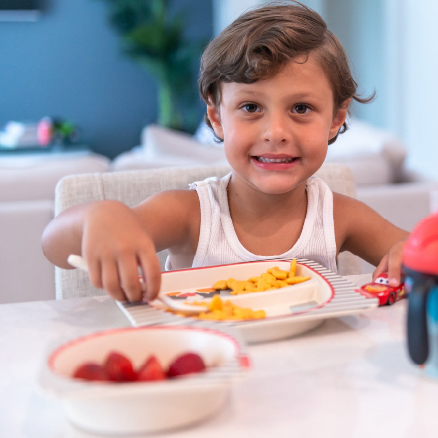 A young child with brown hair sitting in a high chair, smiling at the camera while eating cereal from a plate. A matching bowl containing strawberries is in the foreground, and a small red toy car is on the table.