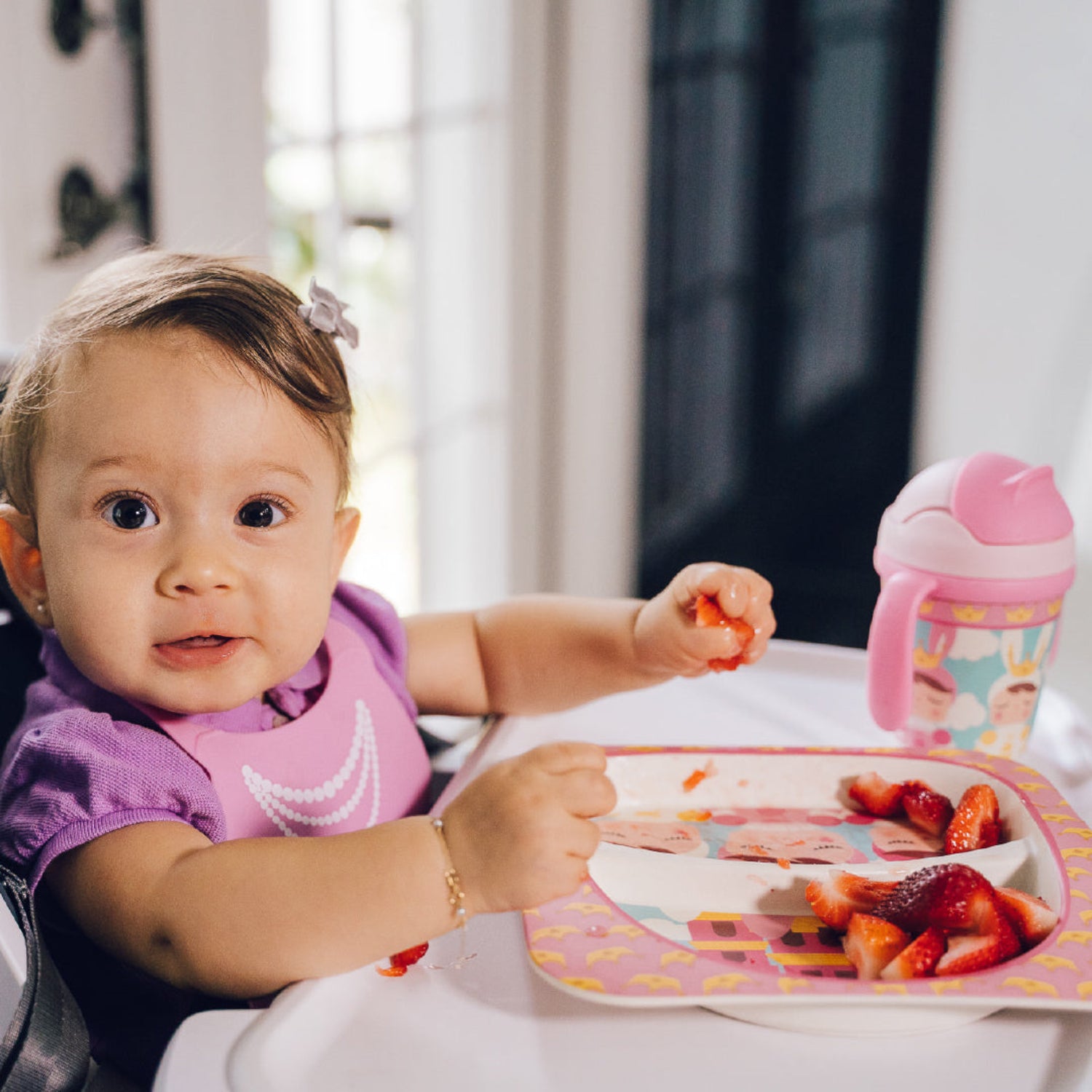 A young child with brown hair sitting in a high chair, eating strawberries from a plate.