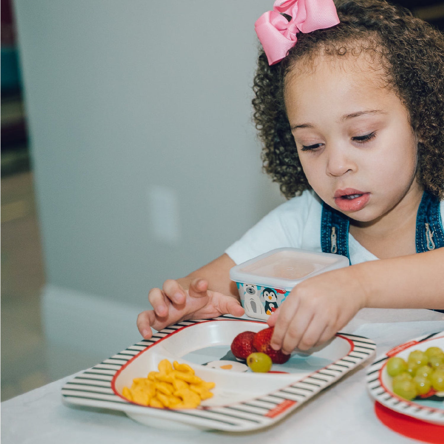 A young girl with brown hair sitting in a  chair, looking at the food while eating strawberry from a plate with a container close to her. A matching bowl containing grapes is in the foreground.