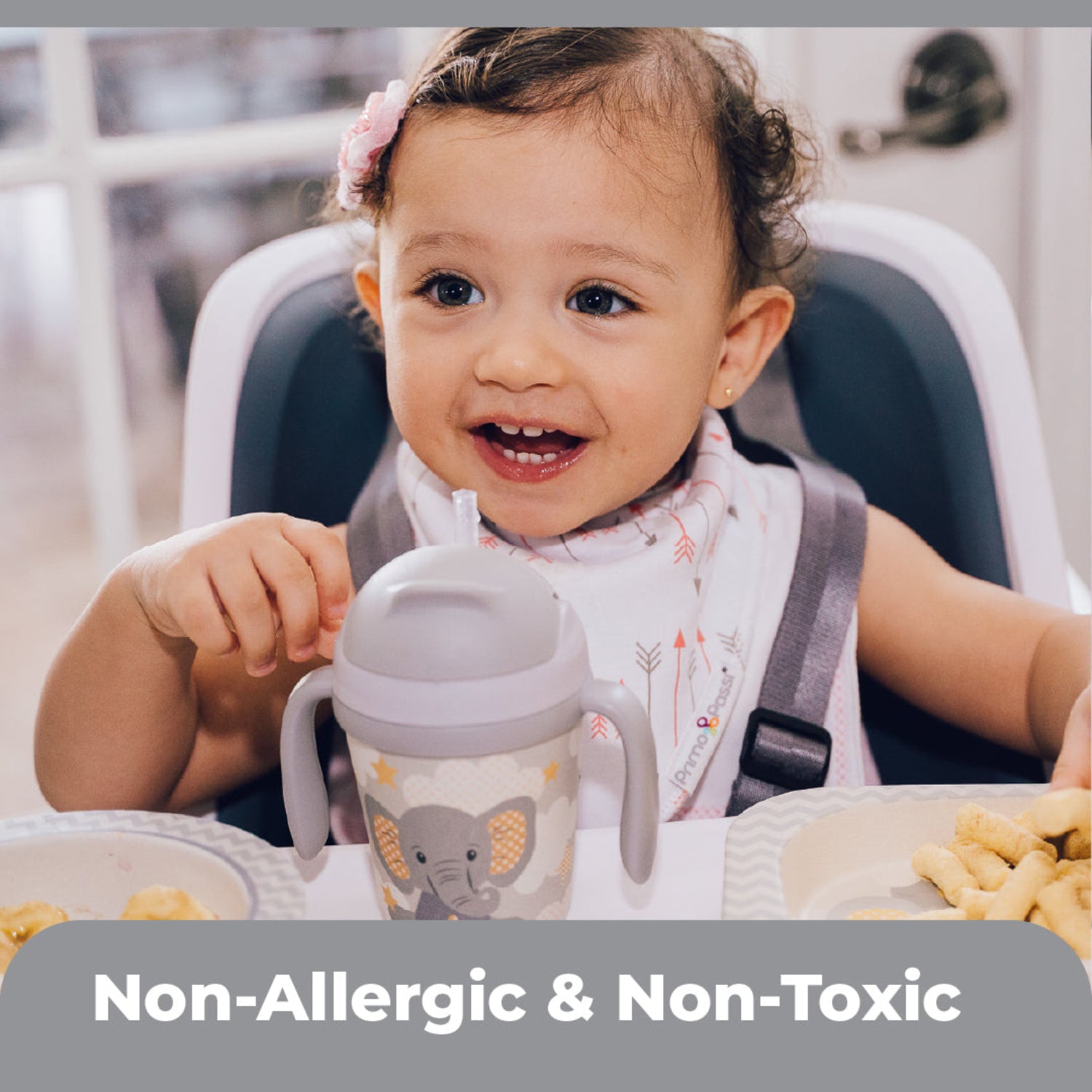 A smiling toddler sits in a high chair during mealtime wearing a Primo Passi white bandana bib with pink and gray arrow prints. The soft, absorbent bib is made with non-allergenic, non-toxic materials to help keep little ones clean and comfortable while eating.