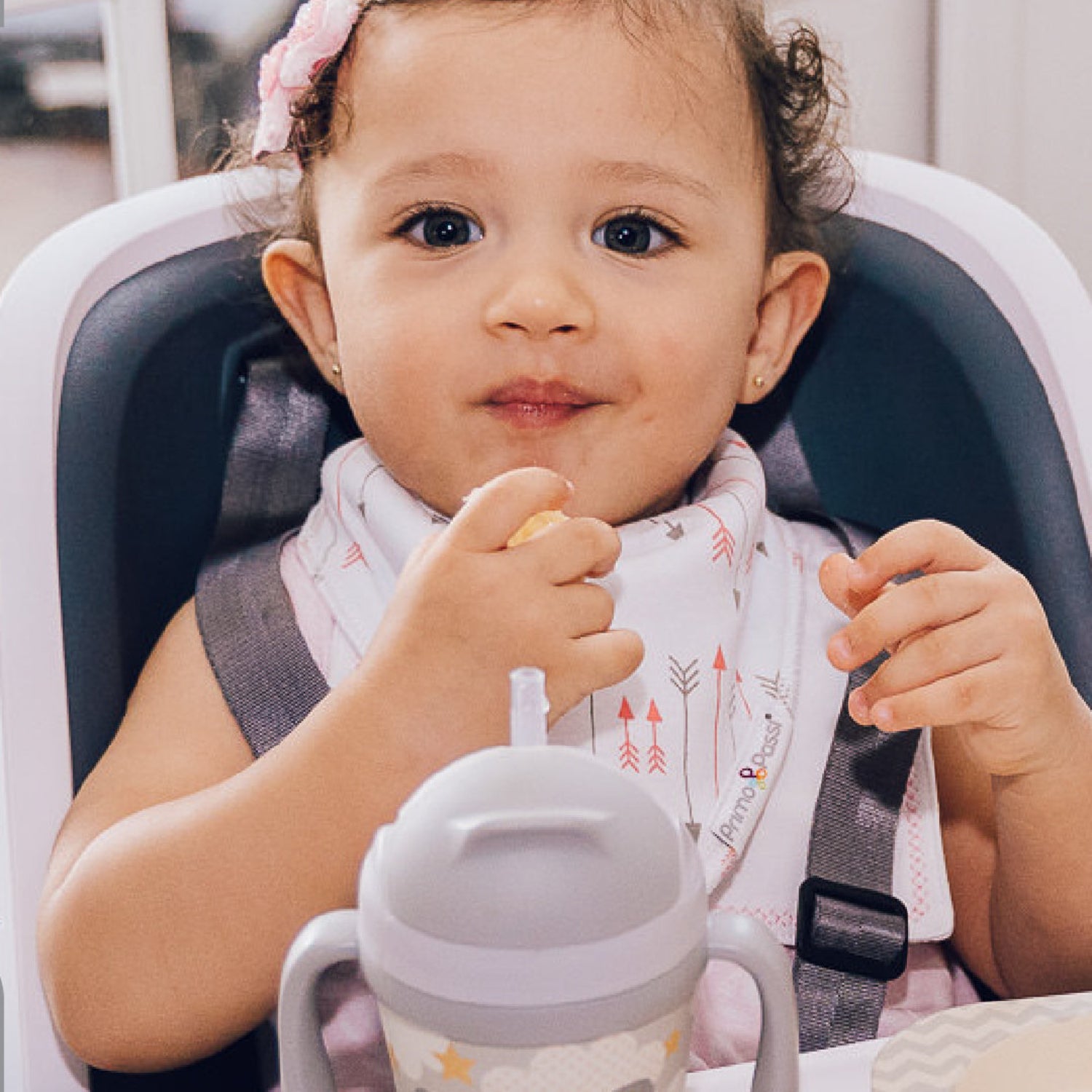 A toddler sits in a high chair enjoying a snack while wearing a Primo Passi white bandana bib with pink and gray arrow prints. The soft, absorbent bib helps protect clothing from drool and mealtime messes while keeping little ones comfortable throughout the day.