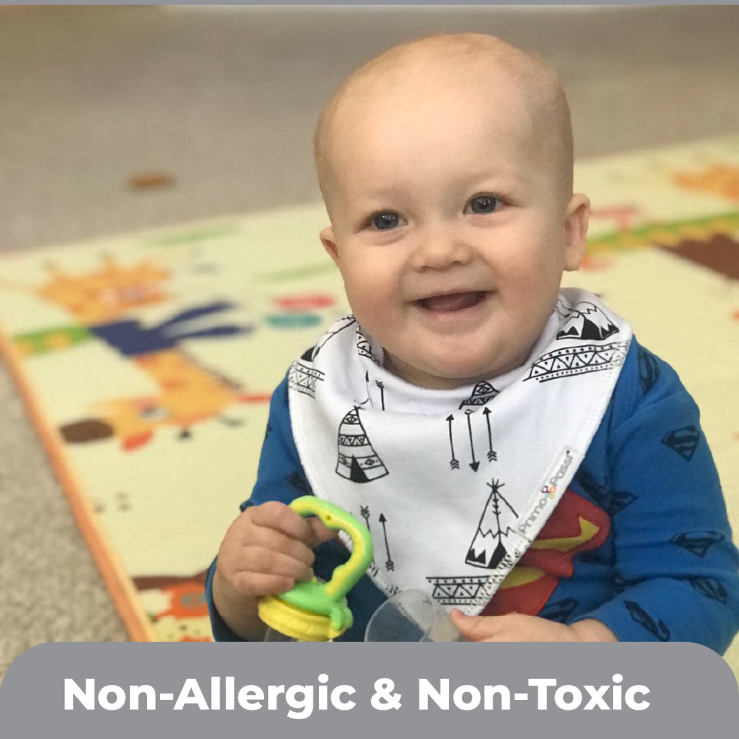 A smiling baby wearing a Primo Passi black-and-white tribal print bandana bib sits on a colorful play mat while holding a teething toy. The soft, absorbent bib is made with non-allergenic, non-toxic materials to help keep babies comfortable during teething and mealtime.