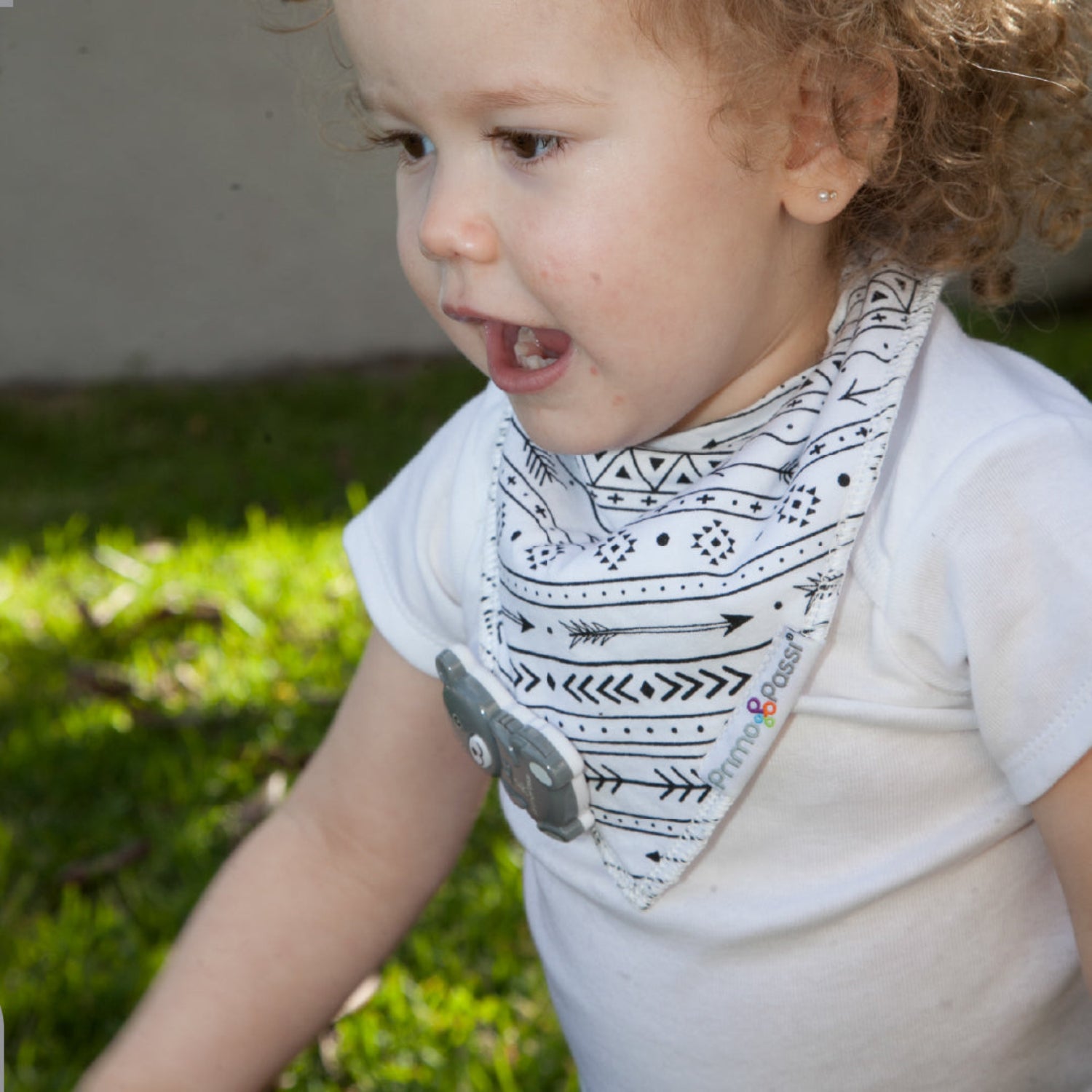 A toddler wearing a Primo Passi black-and-white tribal print bandana bib plays outdoors on the grass. The soft, absorbent bib features a stylish arrow pattern and a secure snap closure, helping keep little ones dry and comfortable during teething, drooling, and everyday adventures.