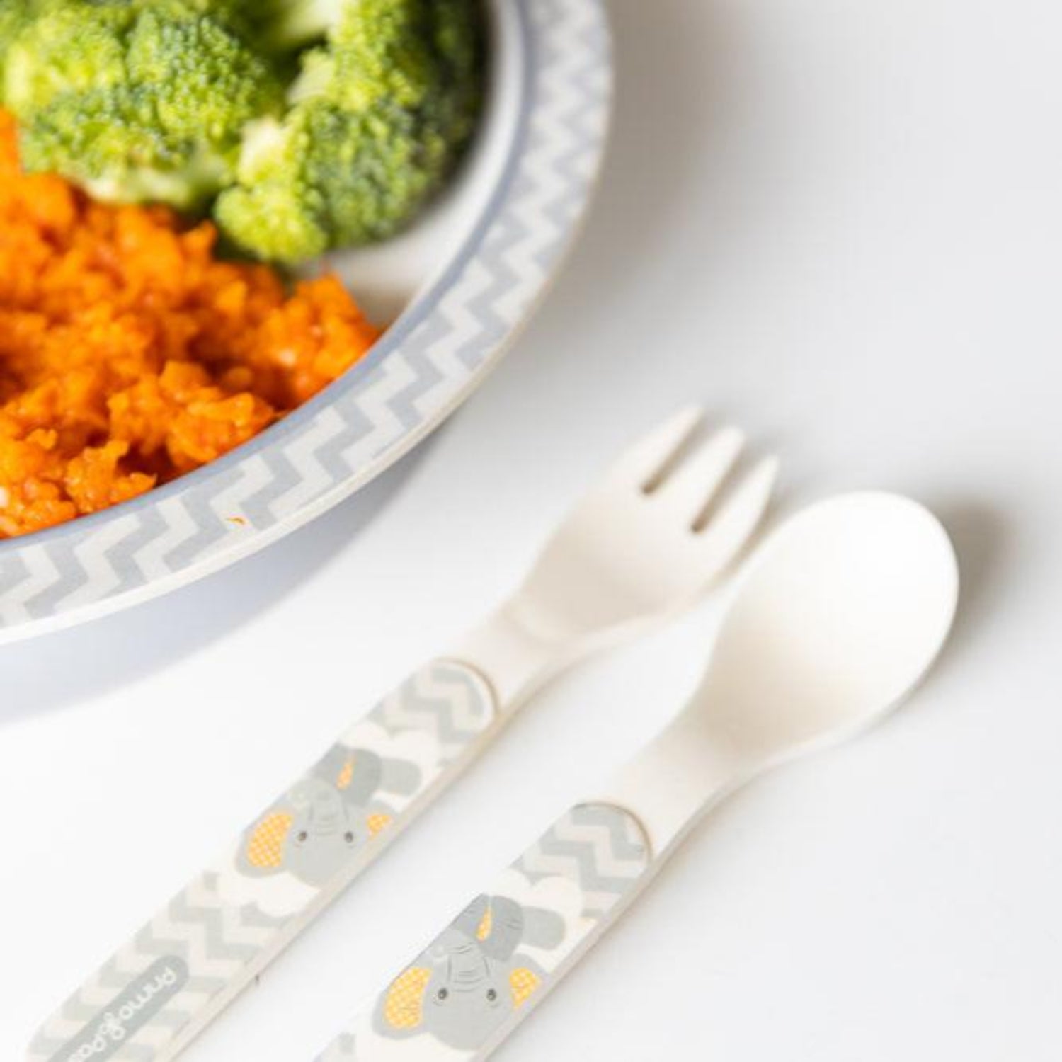 Close-up of a baby spoon and fork set with easy-grip elephant-print handles placed beside a baby plate filled with broccoli and rice. Lightweight utensils are designed to help babies and toddlers learn independent self-feeding safely and comfortably.