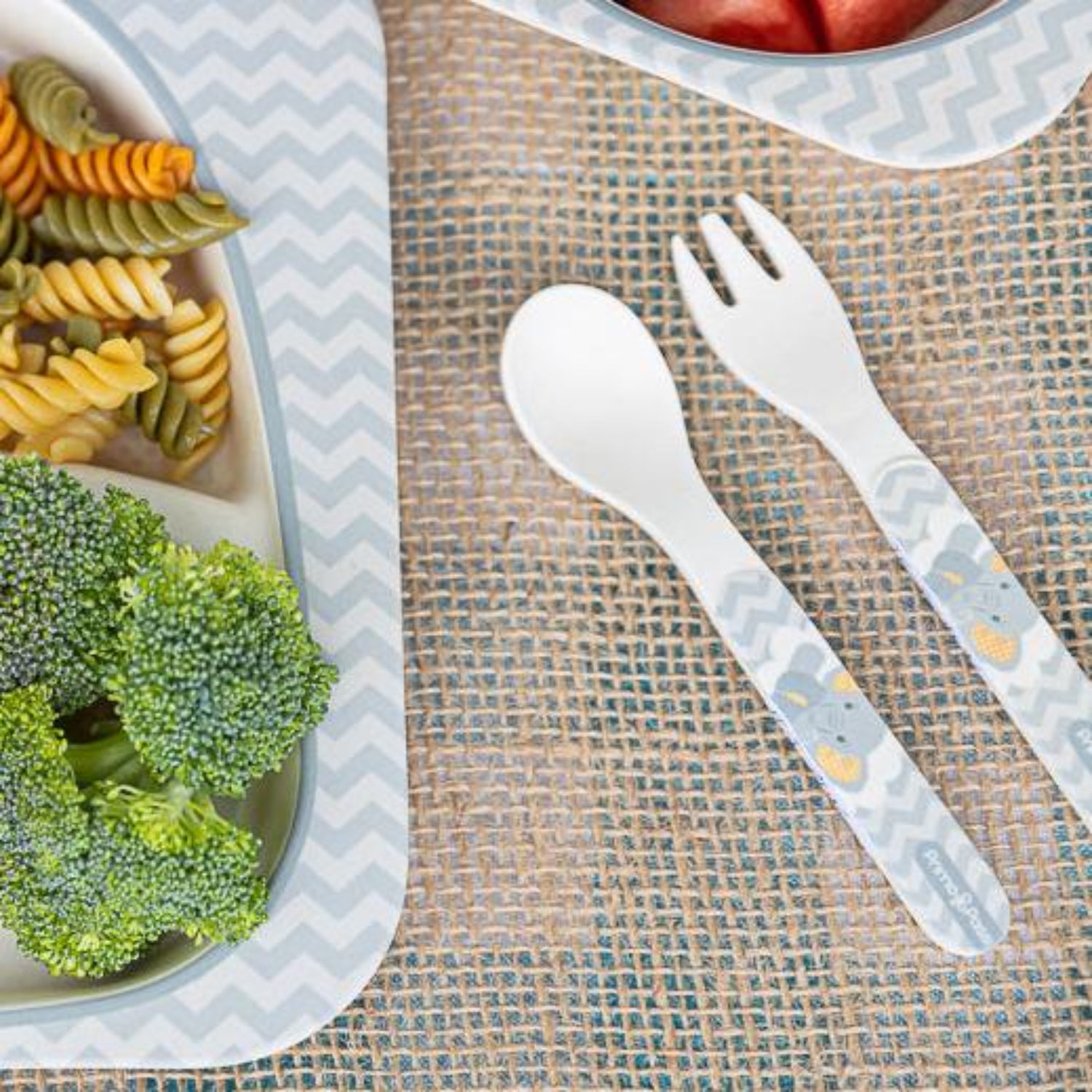 Top view of a baby spoon and fork set with easy-grip elephant-print handles placed beside a divided baby plate filled with broccoli, tri-color pasta, and fresh fruit. Designed to encourage safe, independent self-feeding for babies and toddlers.