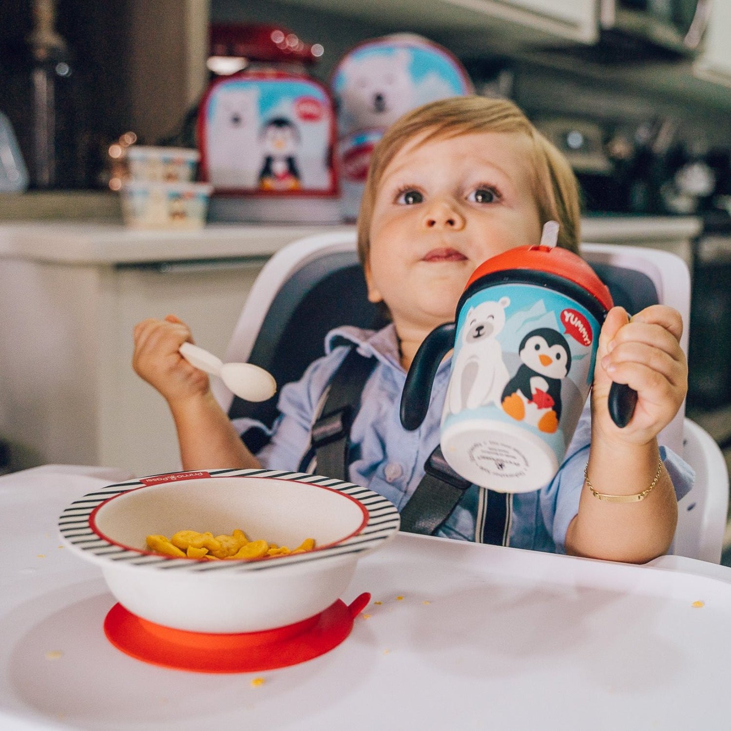 Child in a high chair with a penguin-themed cup and bowl, sitting at a kitchen table.